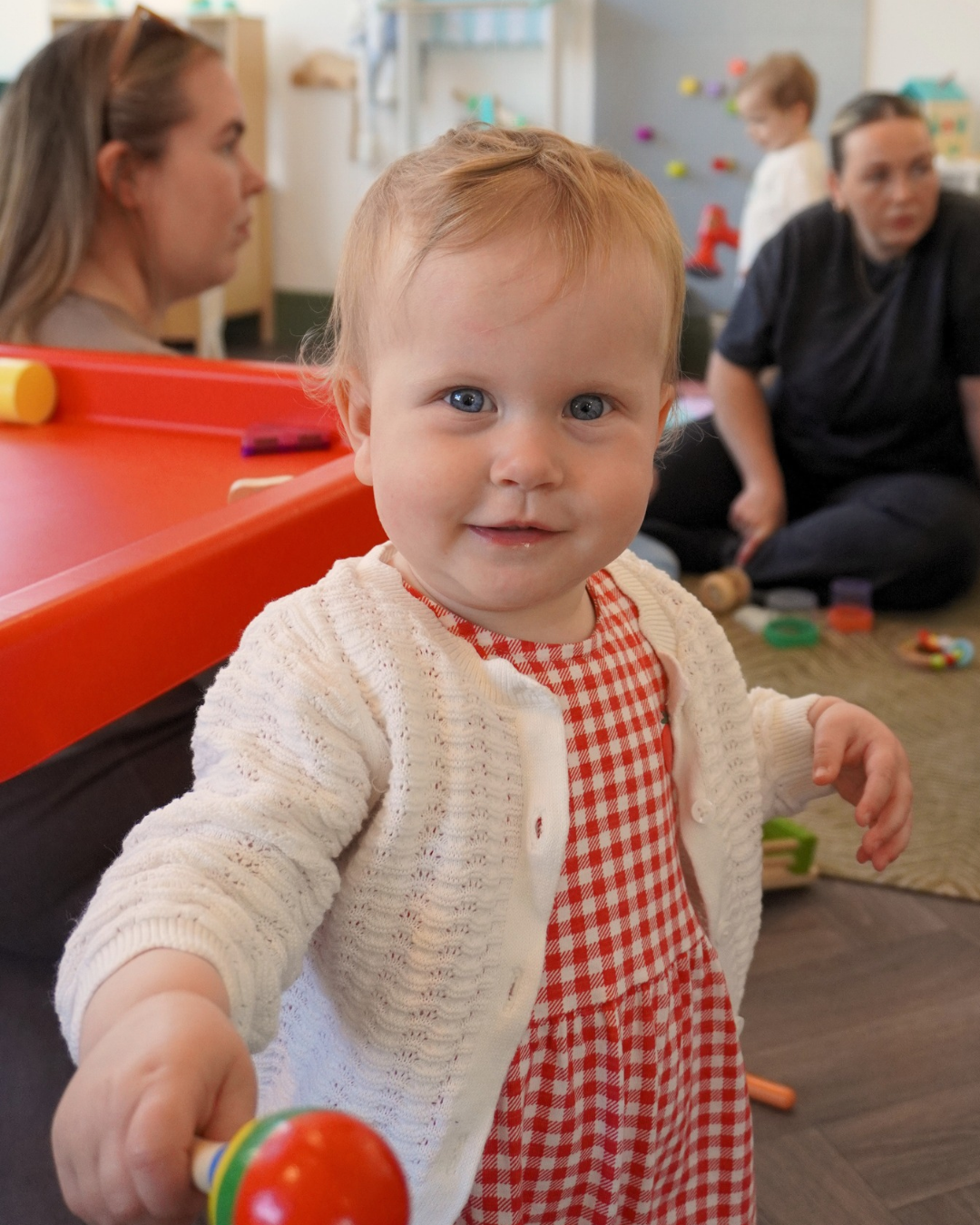 Smiling baby in a white cardigan and red gingham dress holding a colorful toy in a daycare playroom