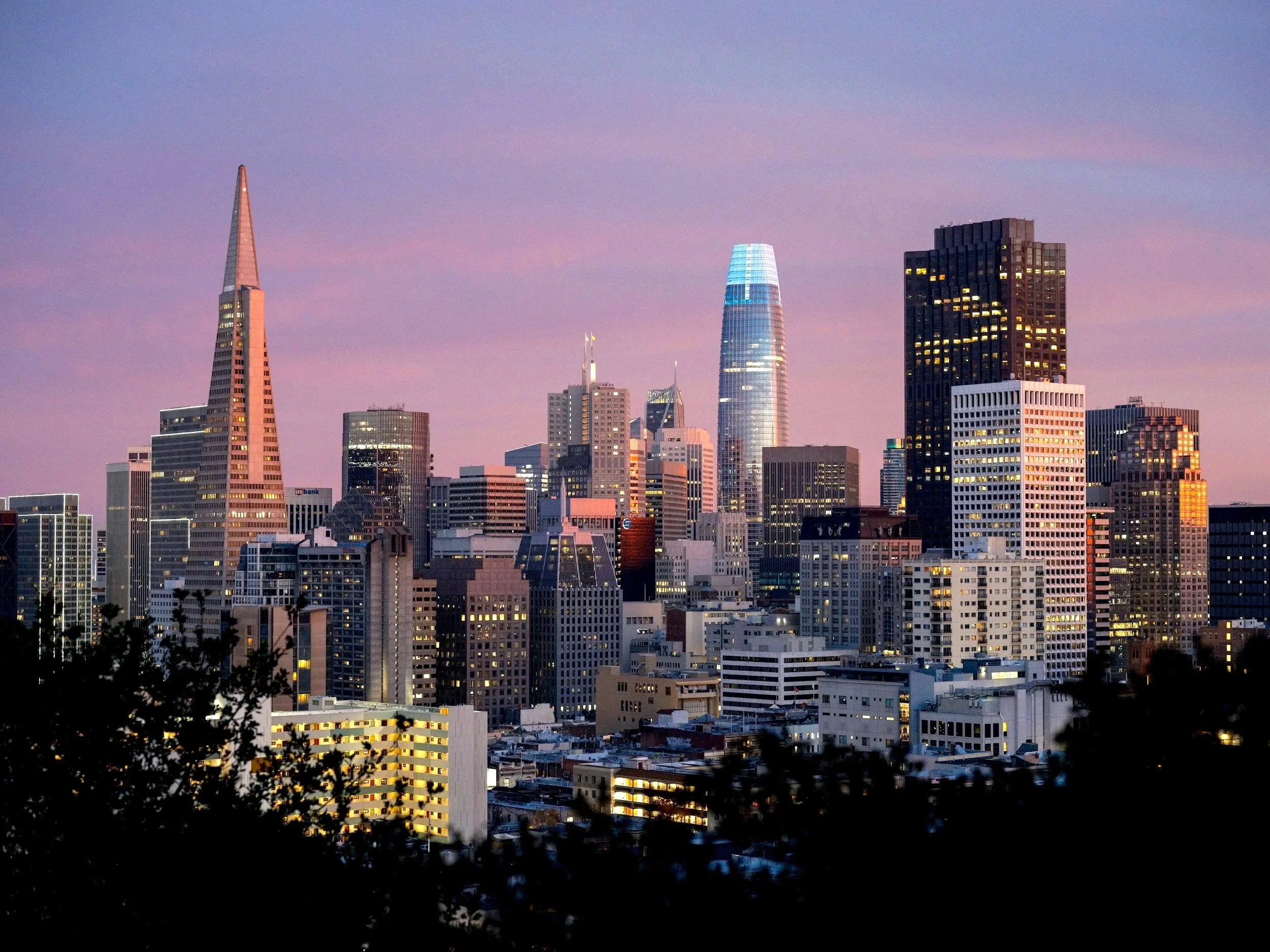 City skyline at sunset with various tall buildings, including the Salesforce Tower with a glass exterior, and the Transamerica Pyramid, set against a purple and pink sky. Construction cranes are visible atop some buildings, and the scene is framed by dark silhouettes of trees in the foreground.
