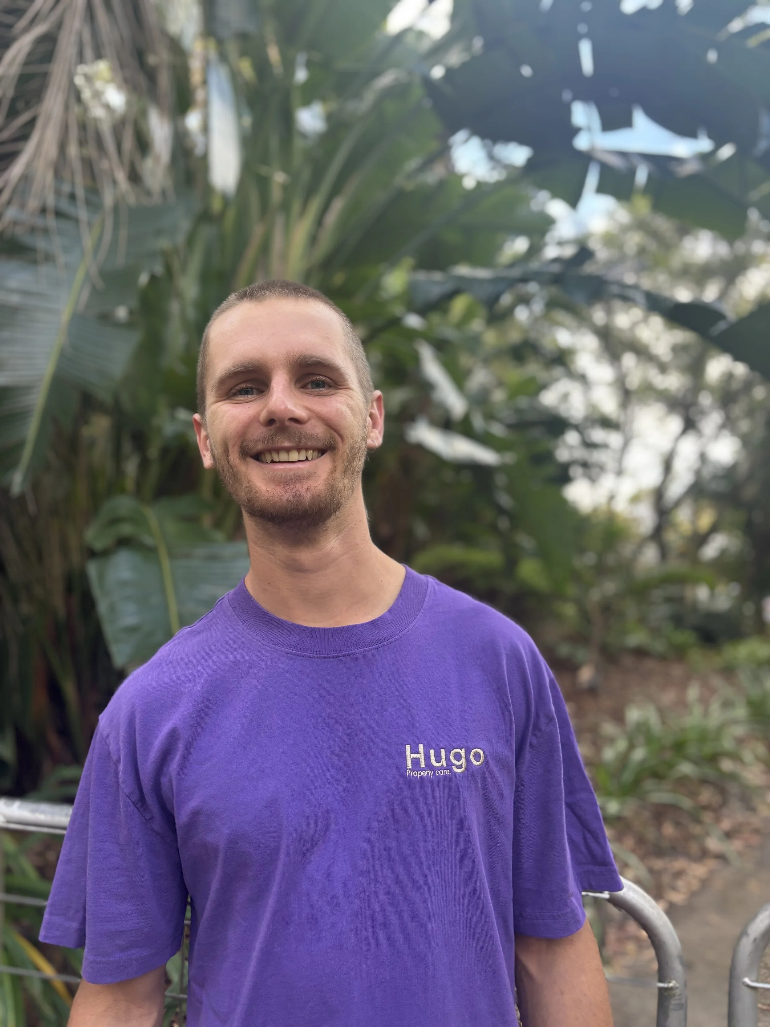 Smiling man wearing a purple 'Hugo Property Care' T-shirt outdoors surrounded by green plants.