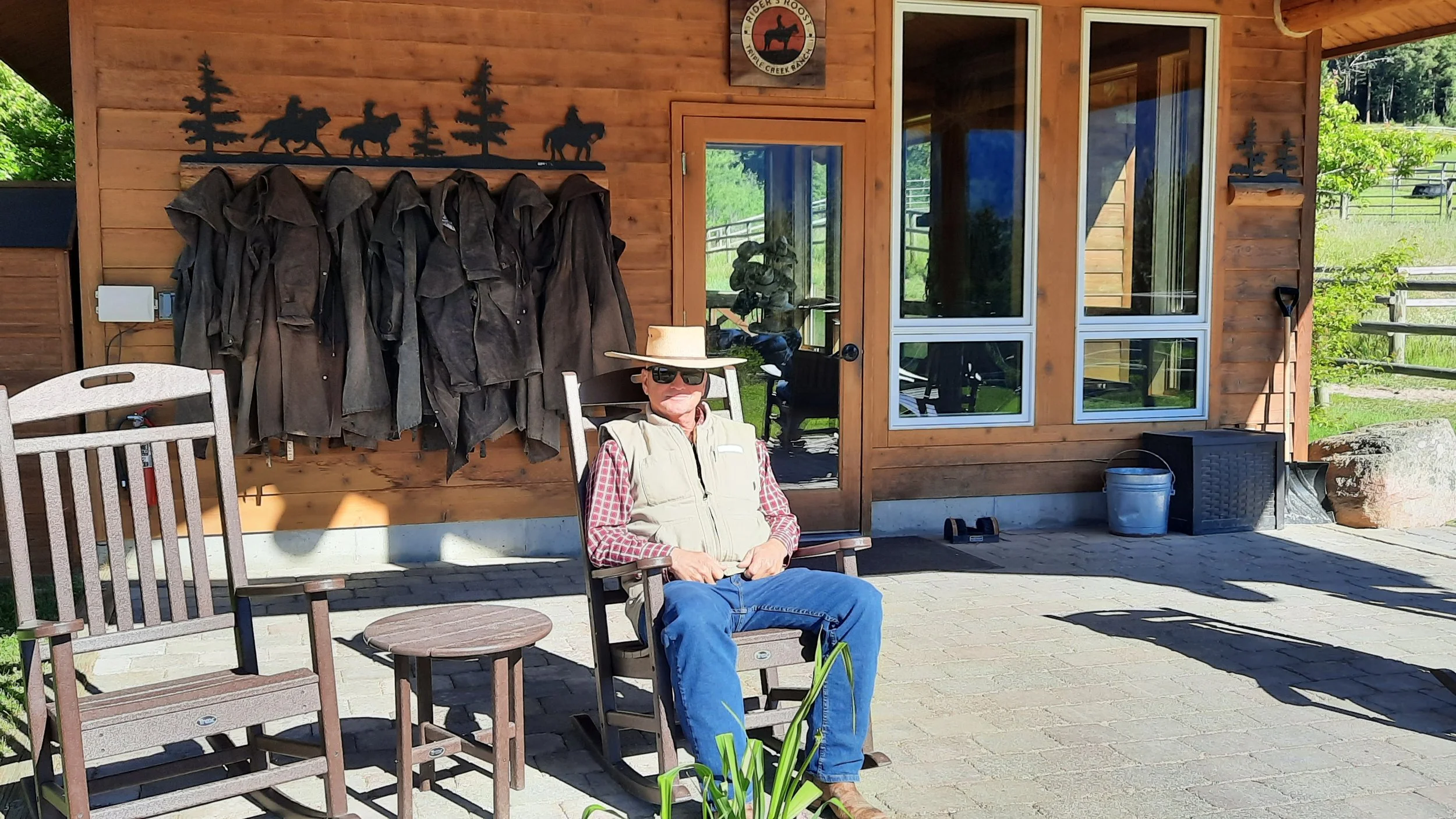 A man wearing sunglasses, a wide-brimmed hat, a red checkered shirt, and a beige vest, sitting outdoors on a wooden chair in front of a cabin with a wood-paneled exterior. Behind him, jackets are hanging on a decorative metal rack featuring silhouettes of trees and horses. The scene is bright with greenery and trees in the background.