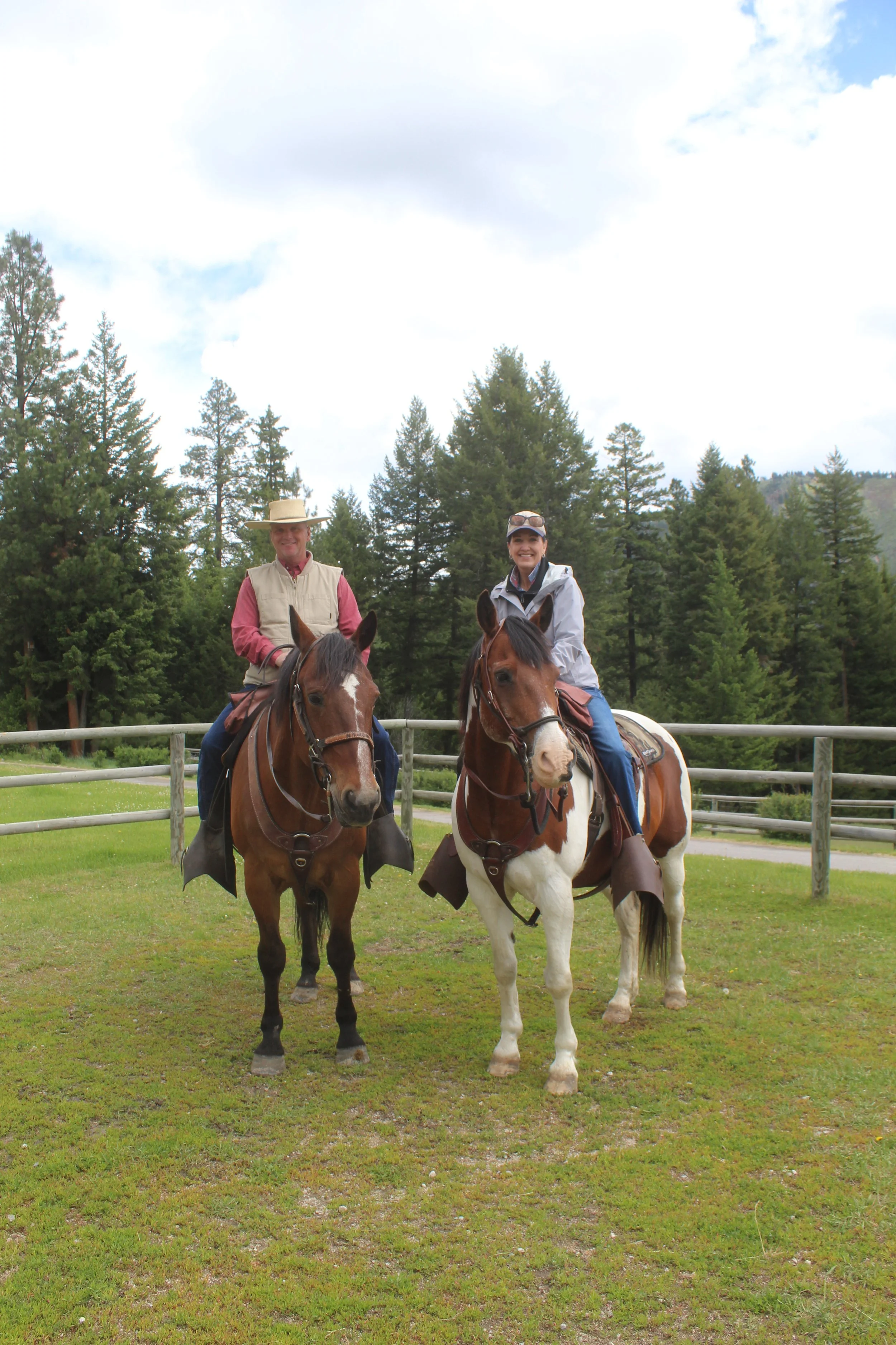 Two people riding horses in a fenced outdoor area with a backdrop of evergreen trees and a partly cloudy sky.