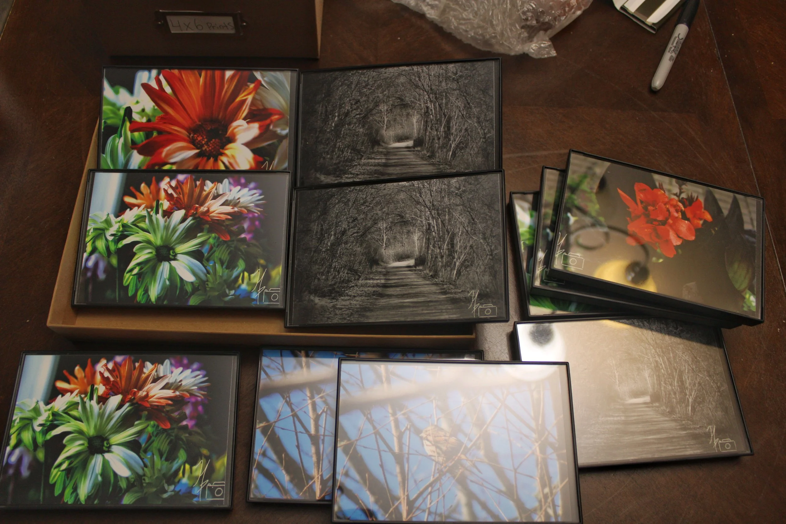 A collection of framed photographs of flowers and nature scenes arranged on a table. The images include colorful flowers and black-and-white scenic views with trees and streams.