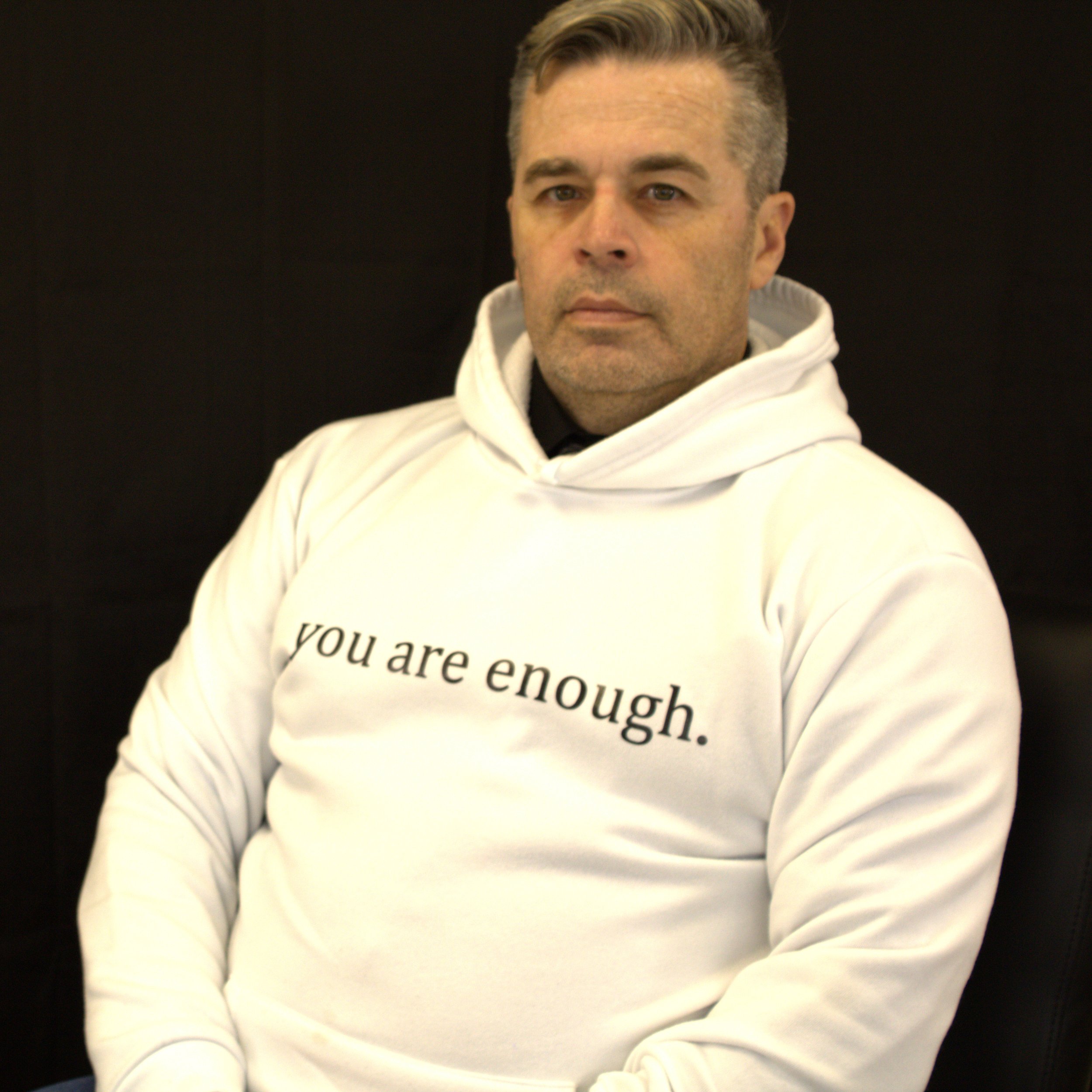 A man with short, styled hair, wearing a white hoodie with partially visible text, sitting against a black background.