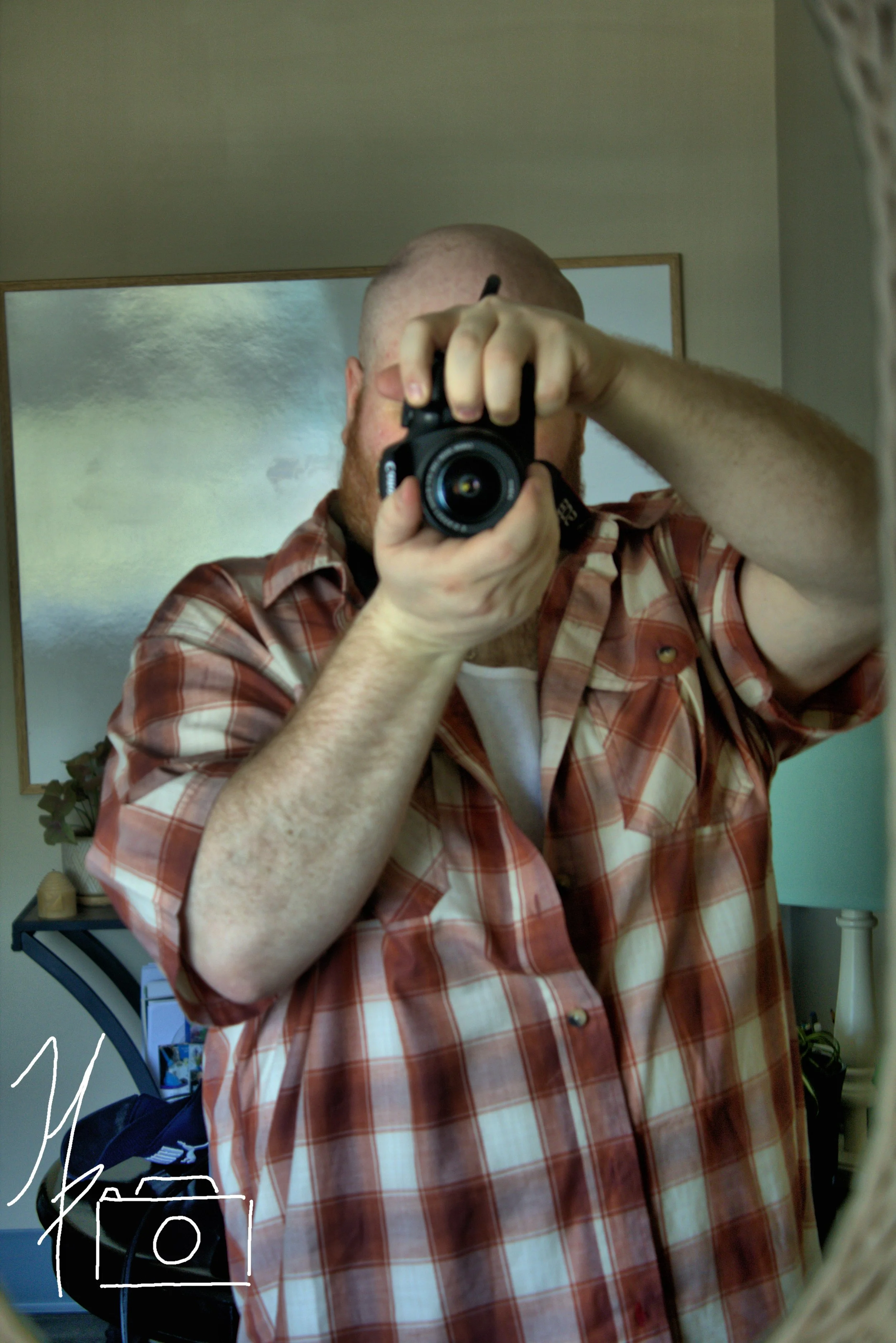 A person taking a selfie in a mirror, wearing a red and white plaid shirt, with a bald head and beard. The person's face is not visible, and the camera obscures their face.