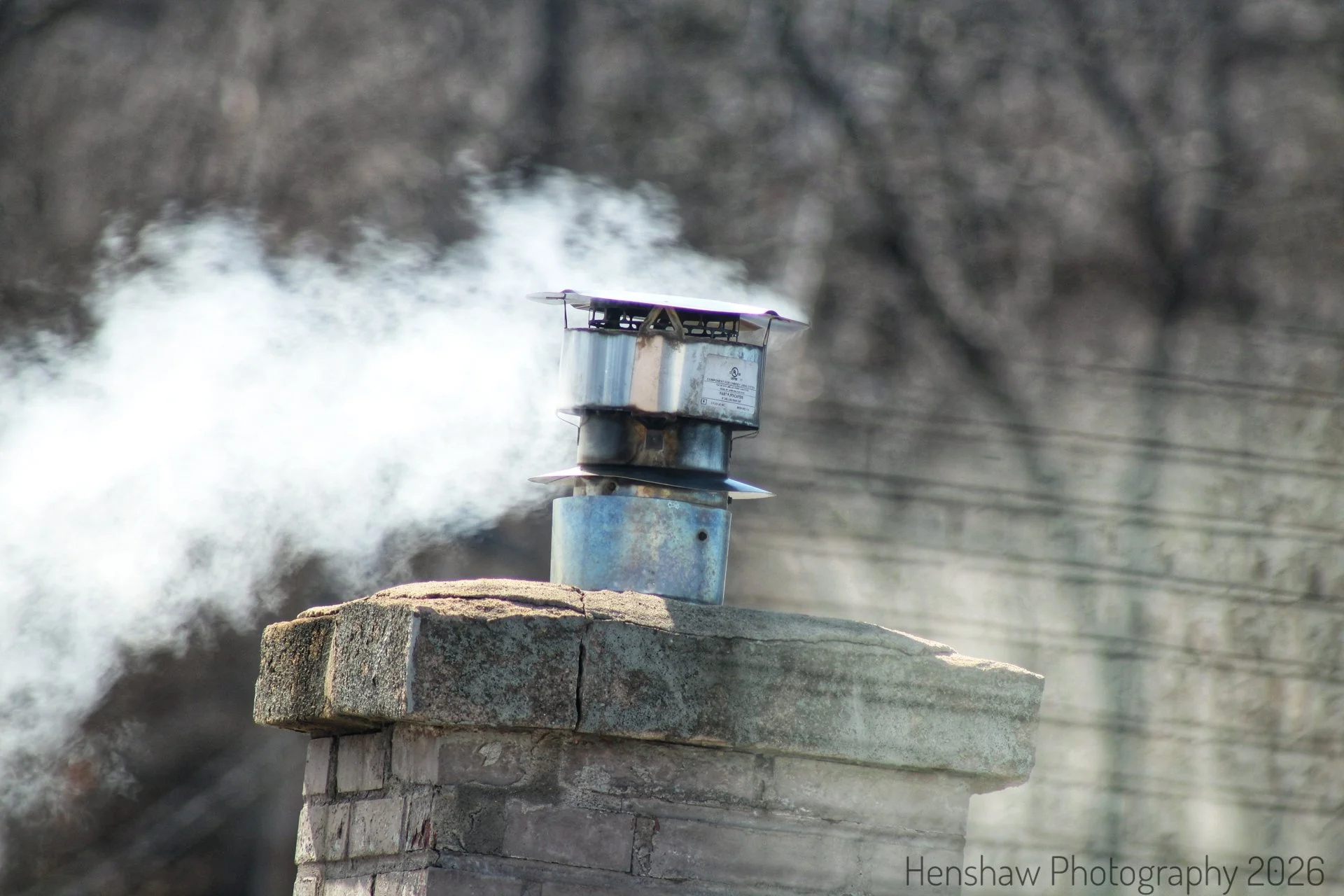 A chimney with steam coming out of it, situated on a brick and concrete ledge. The background shows blurry leafless trees and power lines.
