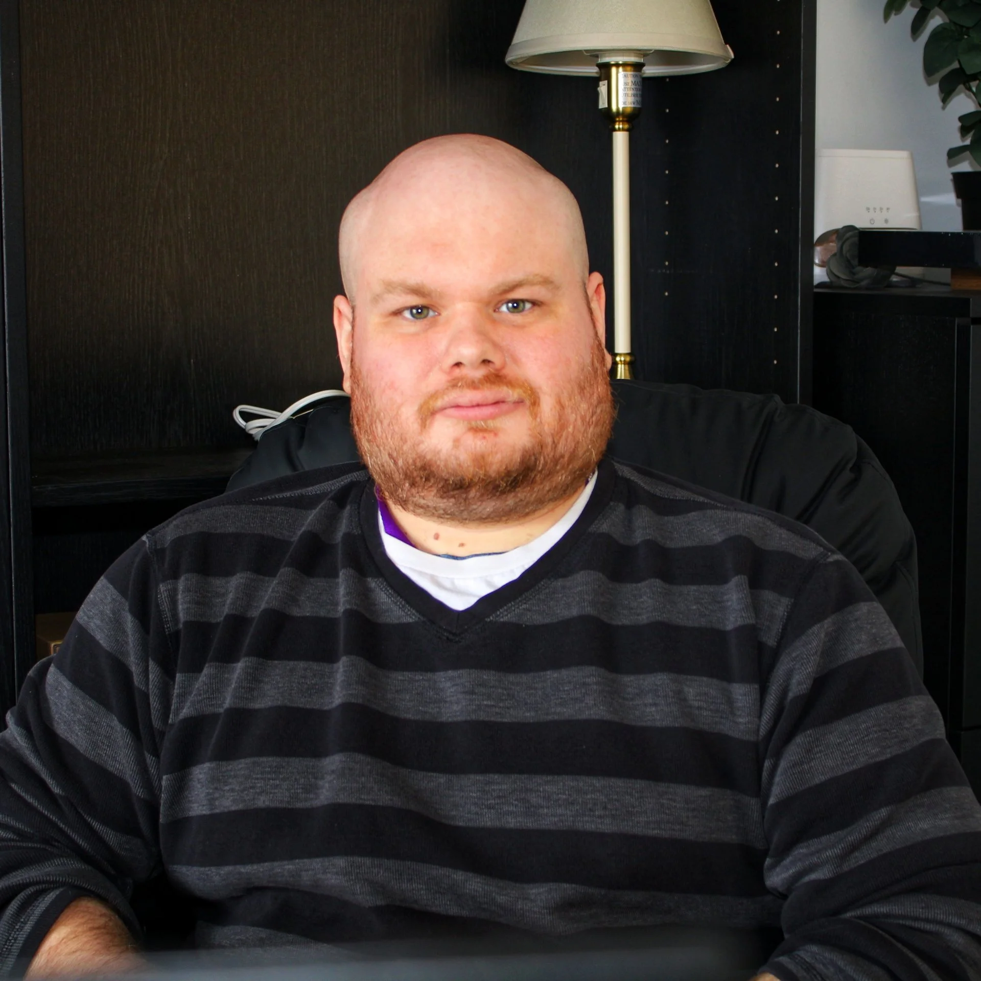 A man with a bald head and a beard sitting indoors, wearing a black and gray striped sweater, with a dark background and a lamp visible behind him.