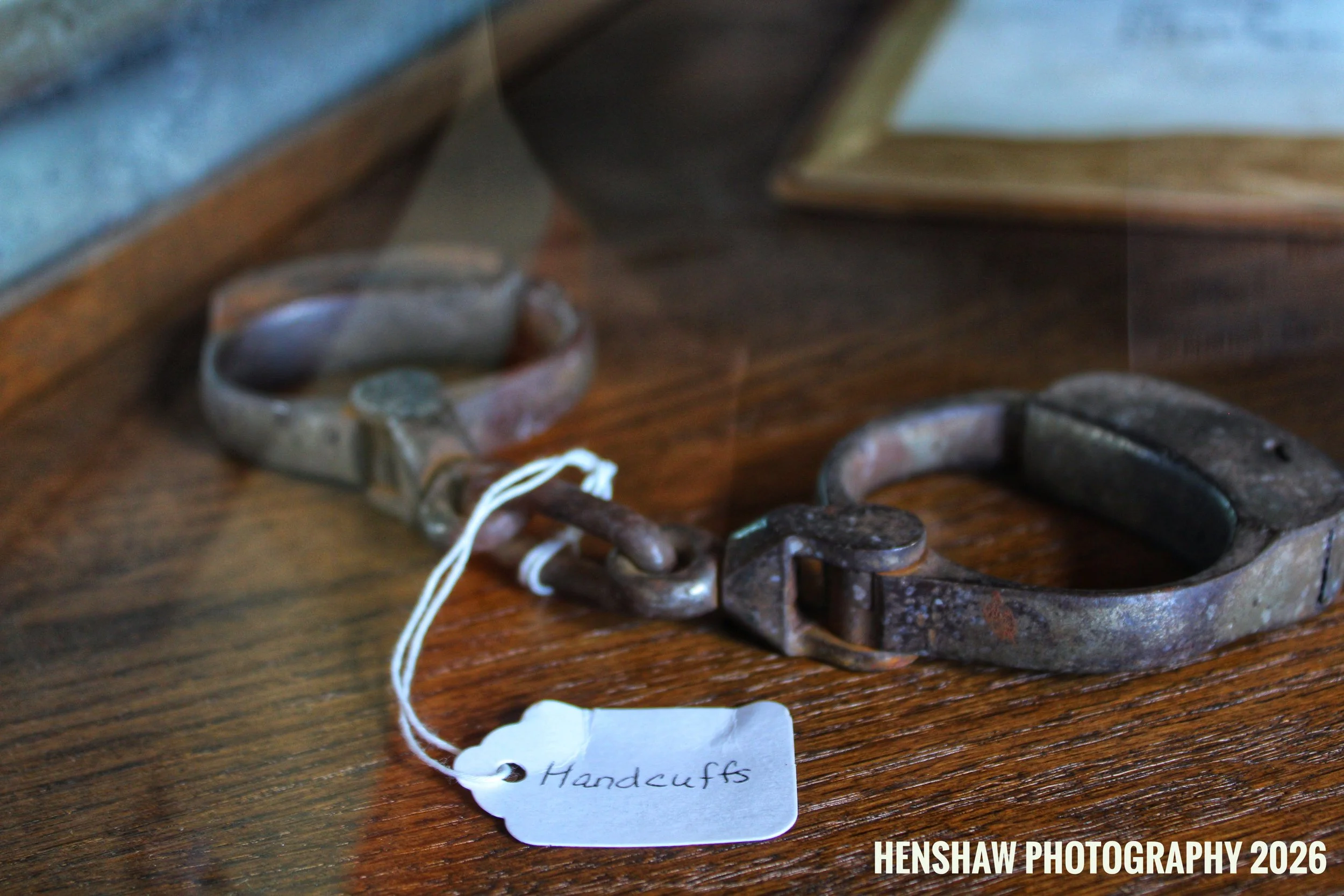 A close-up of a small, rusted handcuff with a white tag attached that reads 'Handcuffs', placed on a wooden surface.