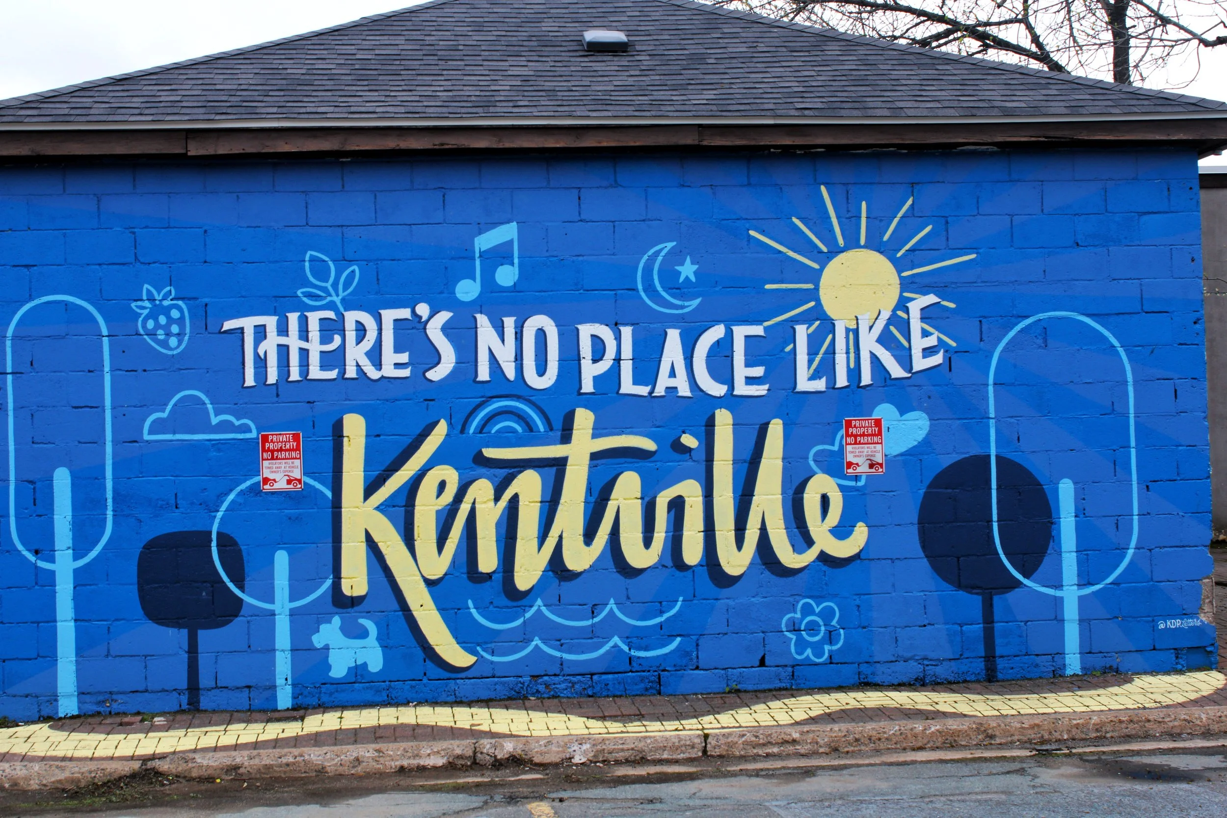 A colorful mural on a blue brick wall with the words 'There's no place like Kentville' written in large, artistic font, surrounded by illustrations of sun, moon, stars, clouds, strawberries, and various whimsical shapes.