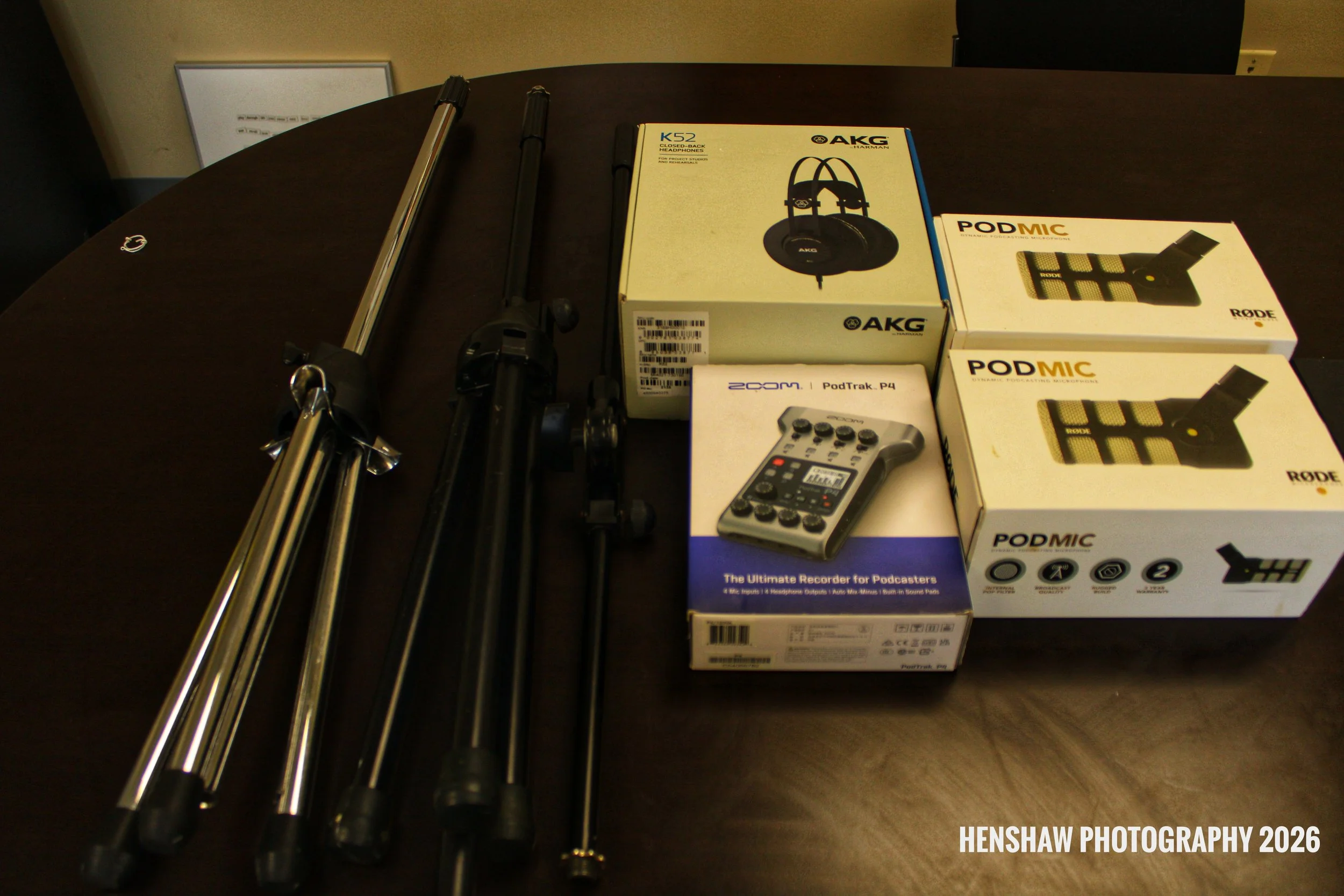 A table with camera tripods, a boxed AKG K52 headphones, Rode PodMic, and Zoom PodTrak P4 podcast recorder.