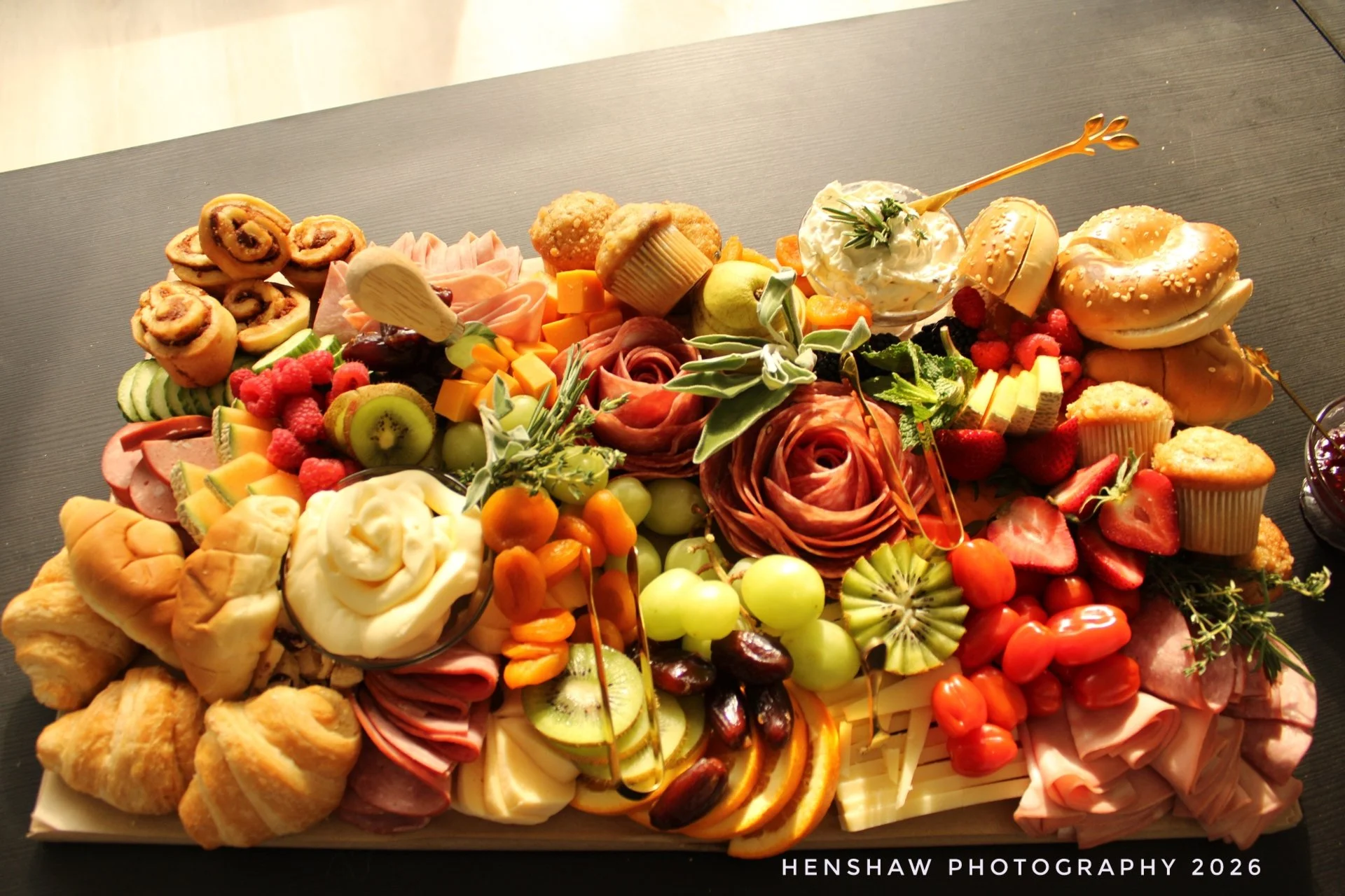 A large tray of assorted finger foods including rolls, fruits like strawberries, kiwi, grapes, and apricots, sliced vegetables, cheese, rolled meats, muffins, and a bowl of dip with a sprig of herbs.