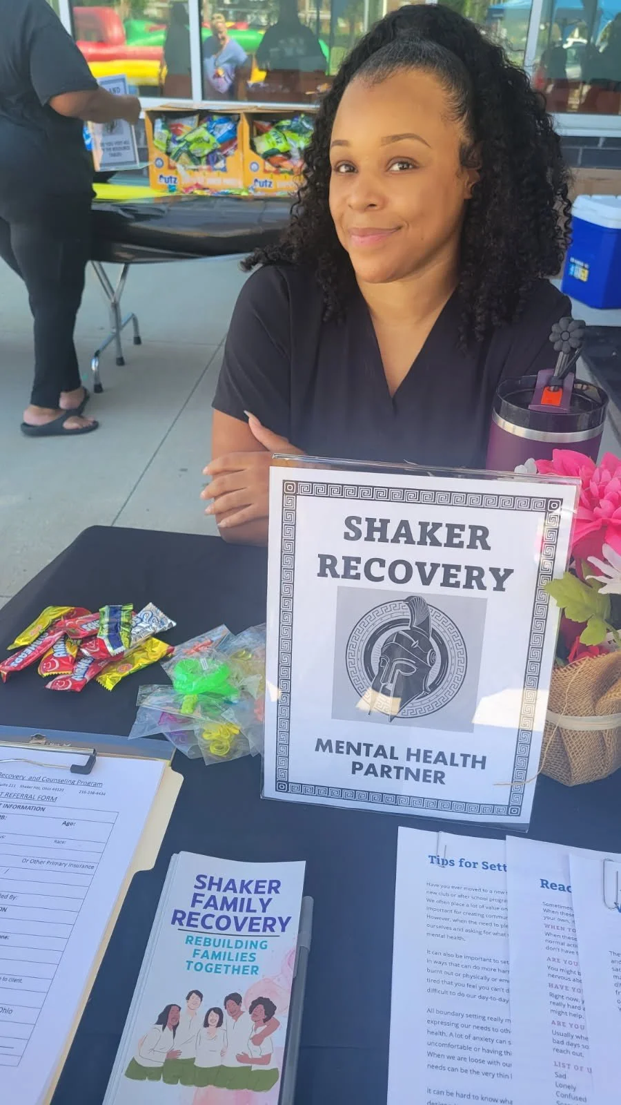 A woman with curly hair sitting at a table with a sign reading 'Shaker Recovery, Mental Health Partner.' The table has assorted candies, flyers, and a pink flower decoration. Behind her are people outside, with a bounce house visible in the background.