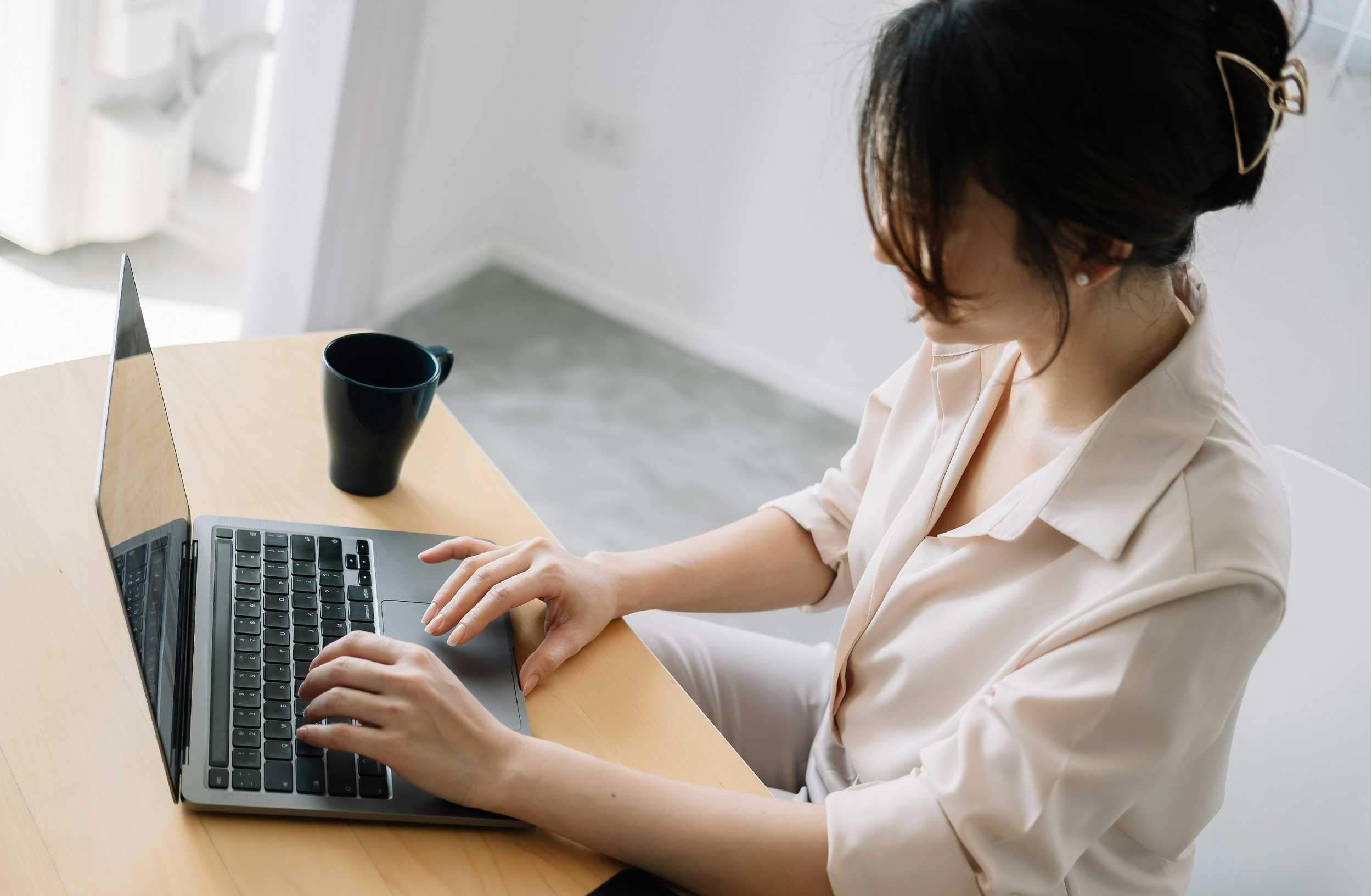 A woman with dark hair secured with a hair accessory, wearing a light beige shirt, working on a laptop at a wooden desk with a black mug nearby in a bright room.