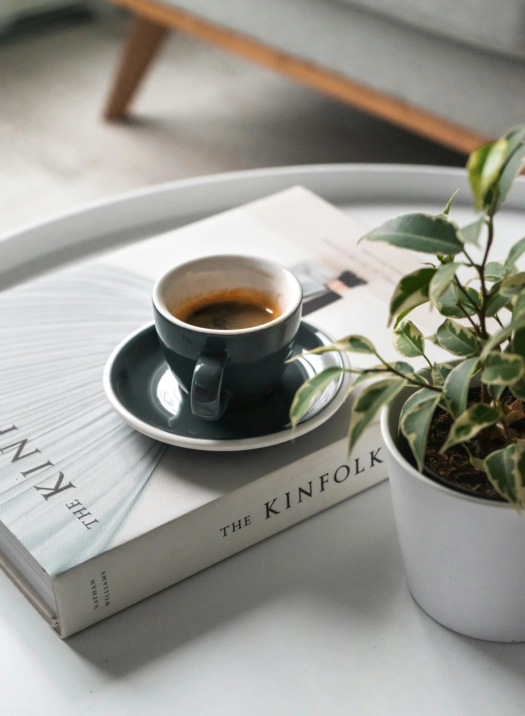 A black coffee cup on a saucer, placed on an open book titled 'The Kinfolk' with a potted plant nearby, all on a white surface.