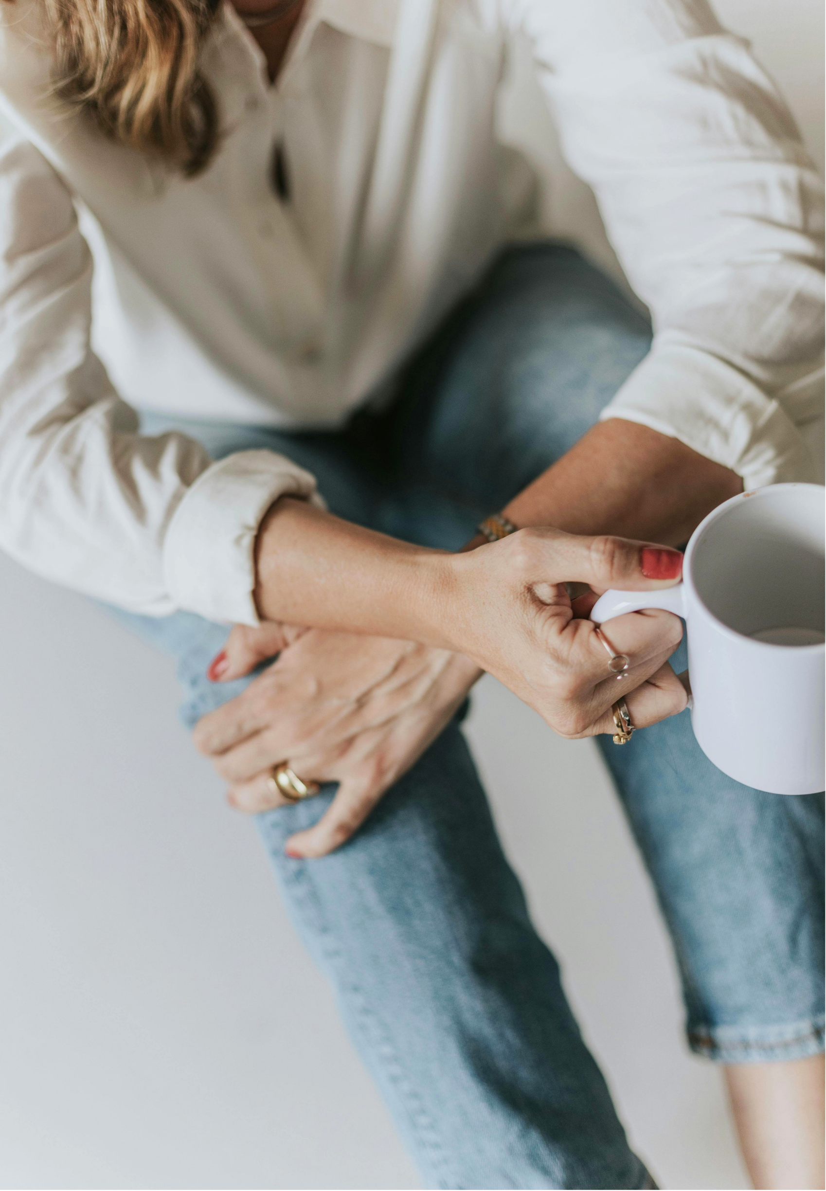 Two women sitting close, one holding a white mug while the other rests her hand on her leg, both wearing casual jeans and a cream-colored shirt.