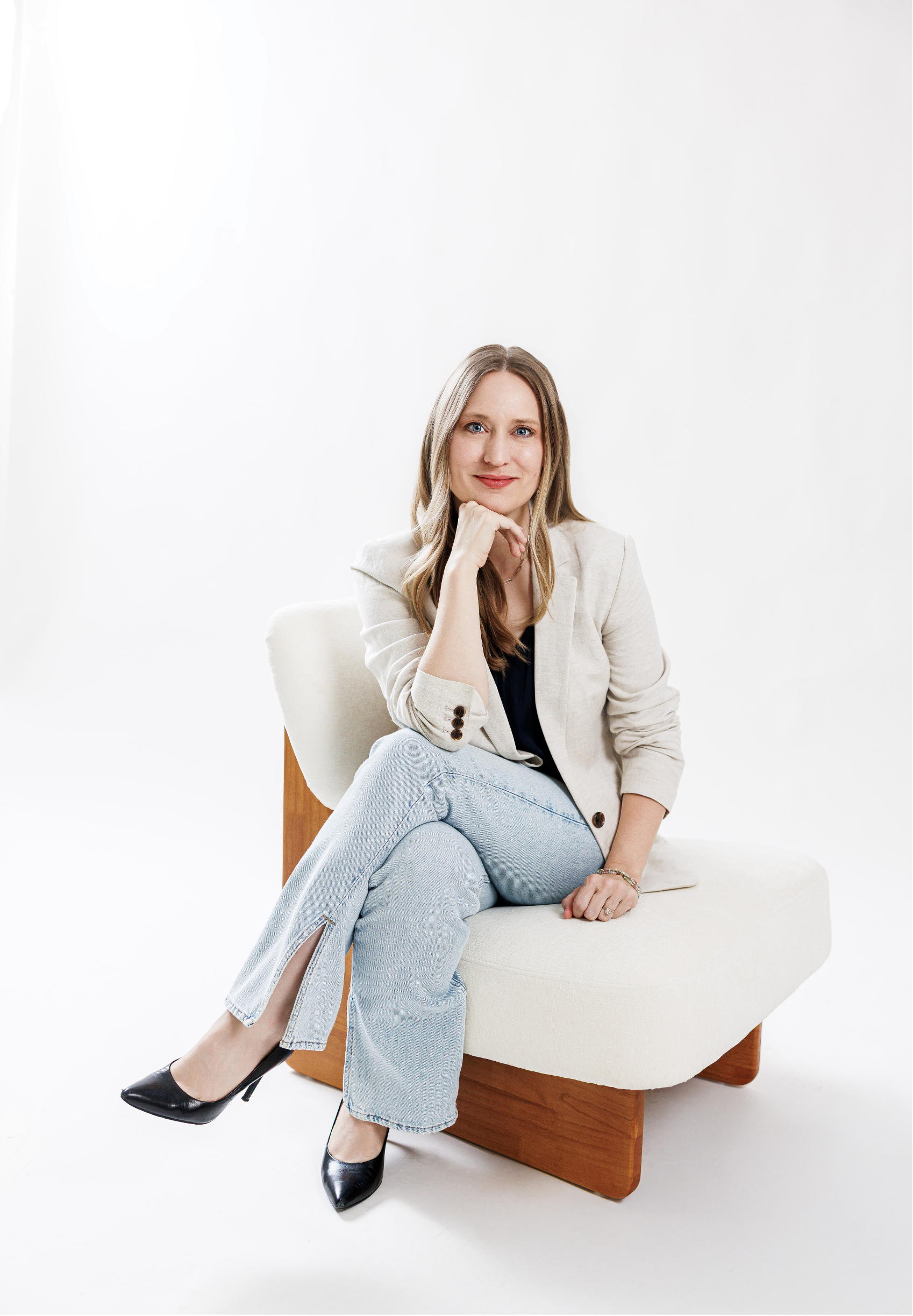 Dr. Laura McKee, Christian counseling in Plano, TX, with blonde hair and blue eyes sitting on a modern white chair with wooden legs, wearing a beige blazer, dark top, light blue jeans, and black high heels, against a plain white background.