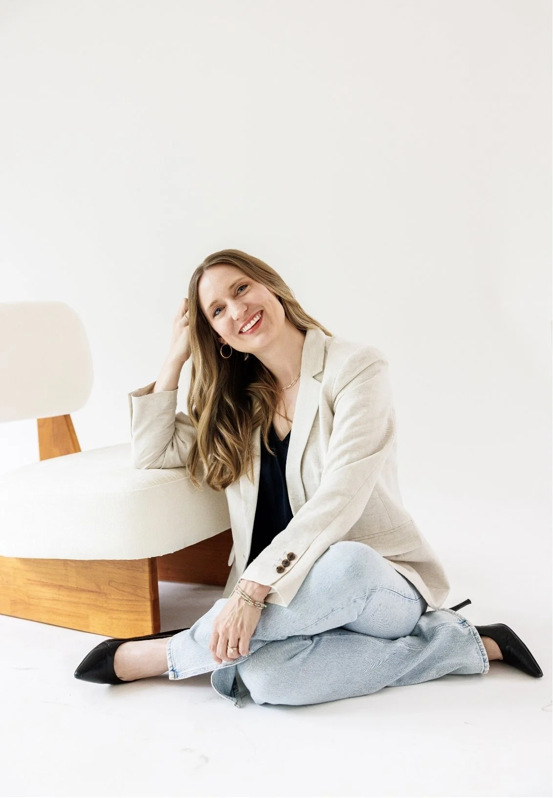 Dr. Laura McKee, Christian counseling in Plano, TX, with long wavy hair, sitting on the floor, smiling, wearing a beige blazer, navy top, light jeans, and black shoes, against a plain white background.
