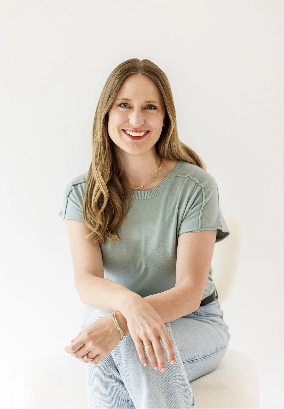 Dr. Laura McKee, Christian counseling in Plano, TX, with long wavy light brown hair, wearing a light green t-shirt, light blue jeans, and jewelry, sitting on a white chair against a plain white background.