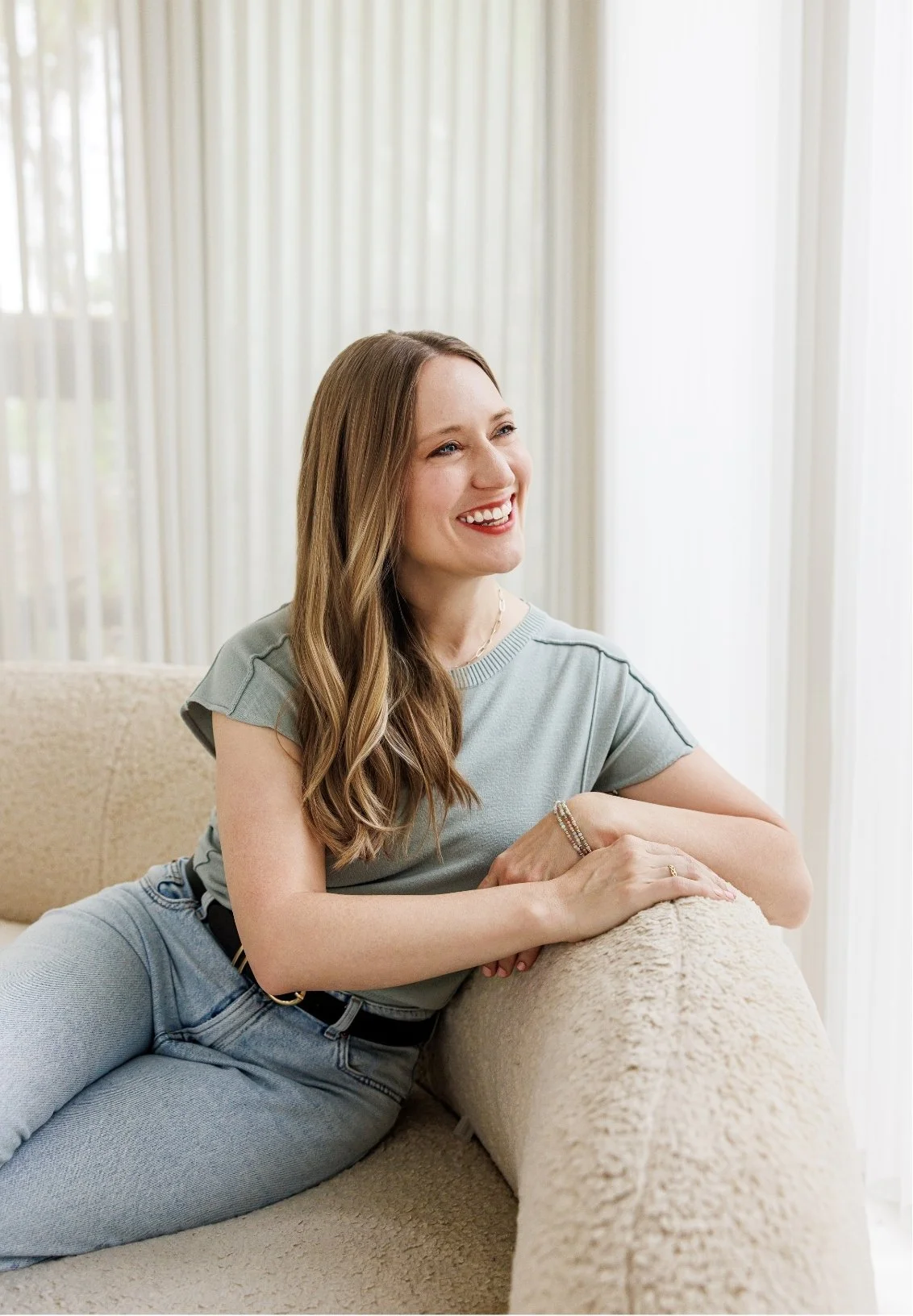 Dr. Laura McKee, Christian counseling in Plano, TX, with long, wavy hair, wearing a light gray t-shirt and jeans, smiling and relaxing on a beige couch in a bright room with light-colored curtains.