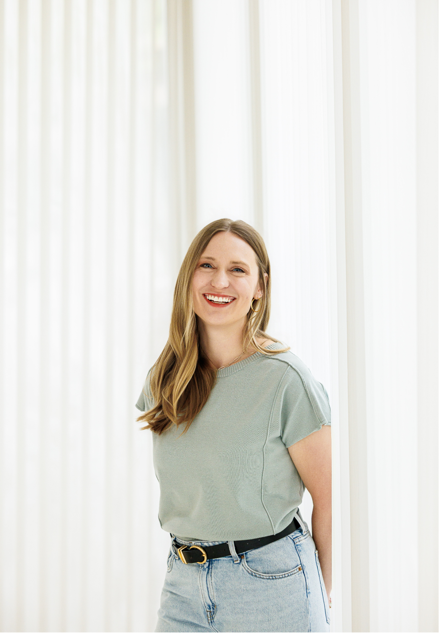 Dr. Laura McKee, Christian counseling in Plano, TX, with long blonde hair wearing a light green T-shirt and light blue jeans standing in front of white curtains.