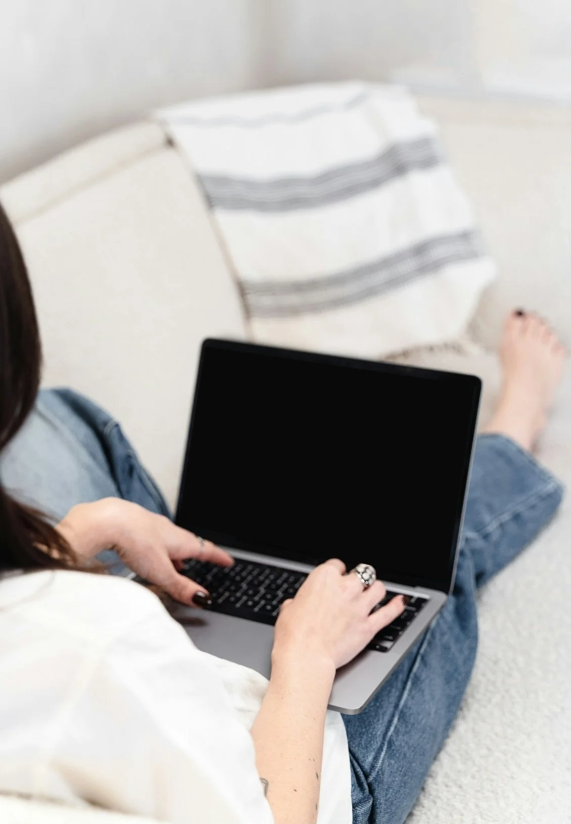 Person sitting on a white couch using a laptop with a black screen, wearing blue jeans and a white shirt, with a striped pillow in the background.