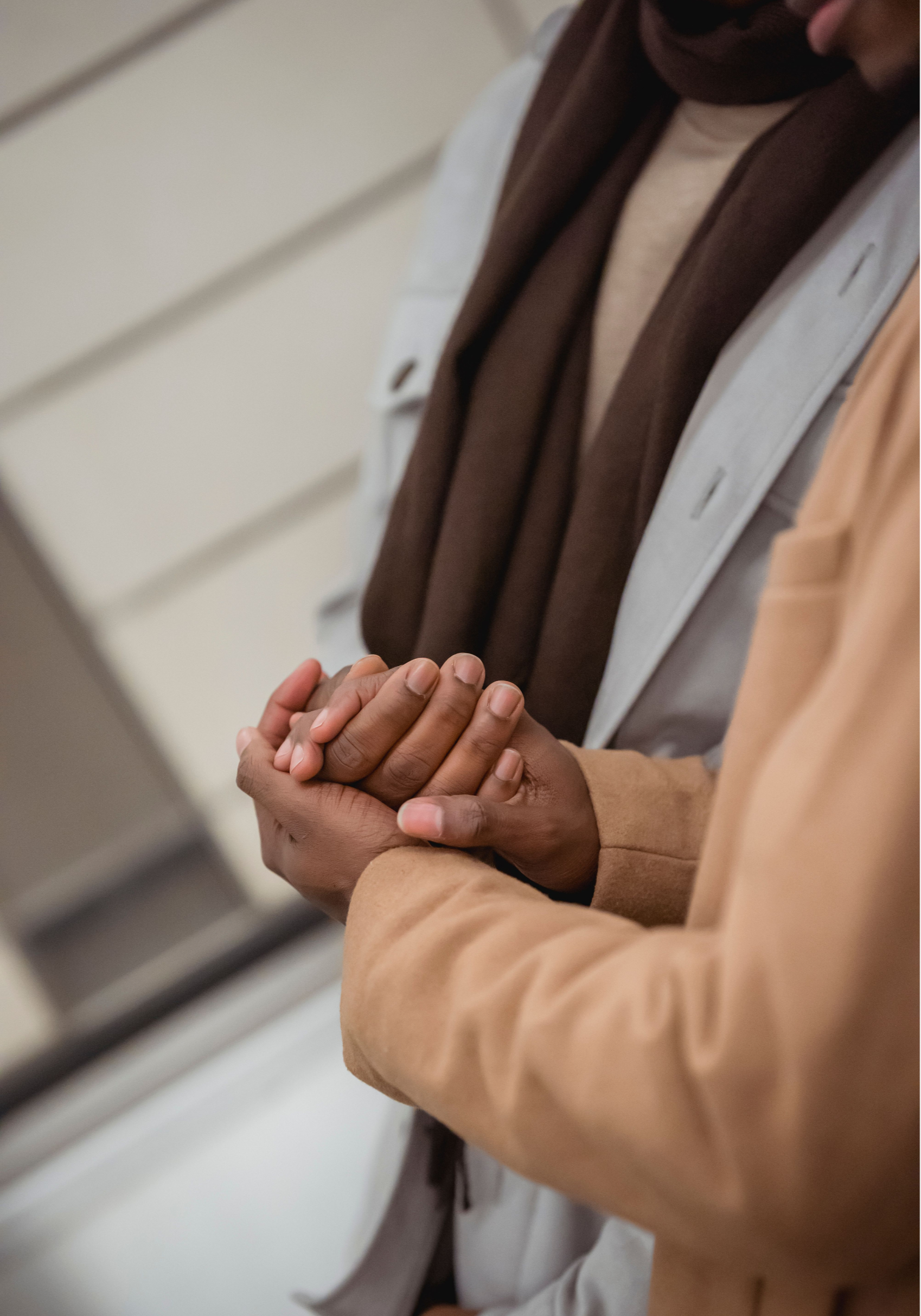Two people holding hands, one with darker skin and the other with lighter skin, standing near beige wall with horizontal slats, wearing brown and beige clothes.