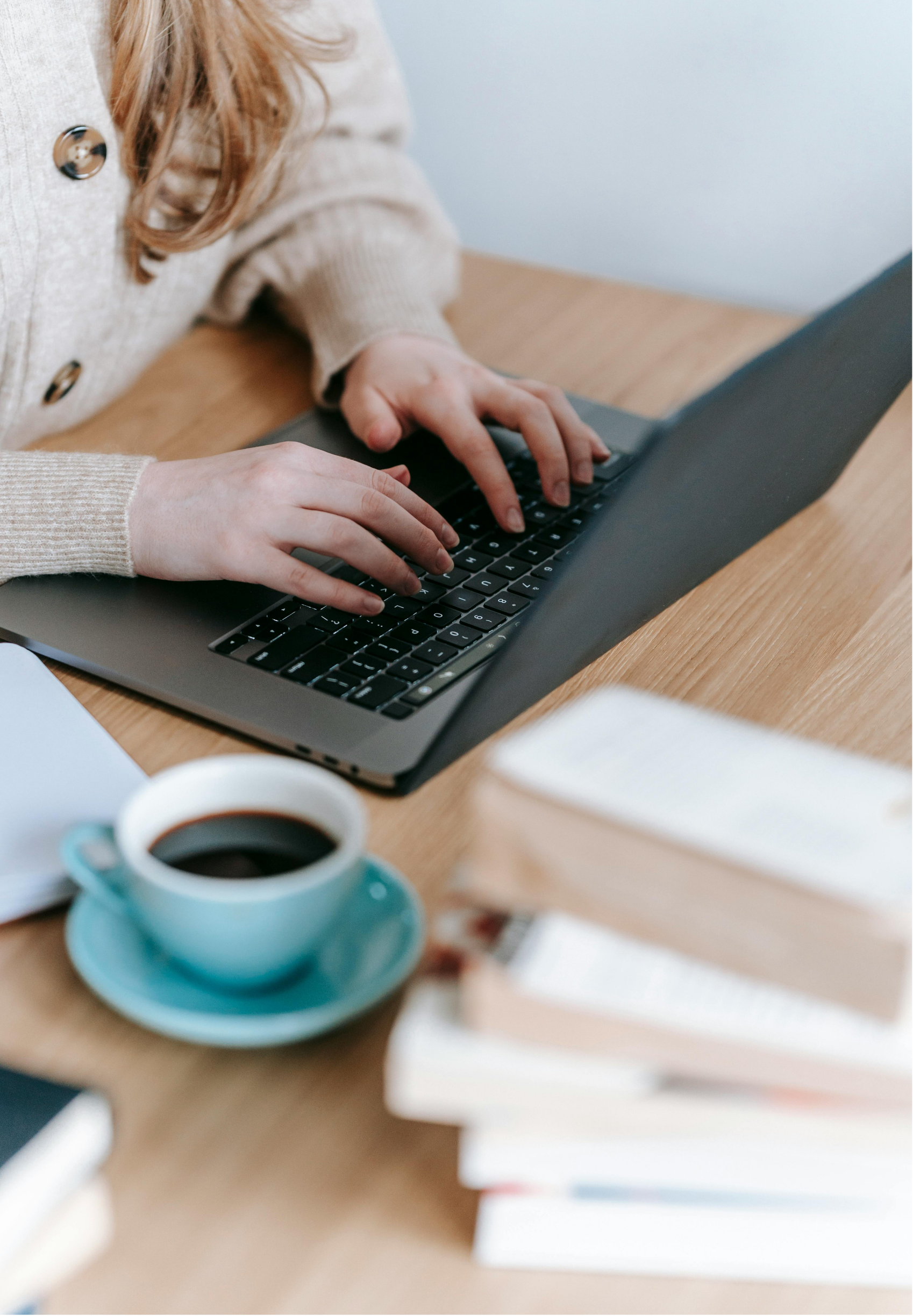 A person wearing a beige sweater typing on a laptop keyboard on a wooden desk, with a cup of coffee and books nearby.