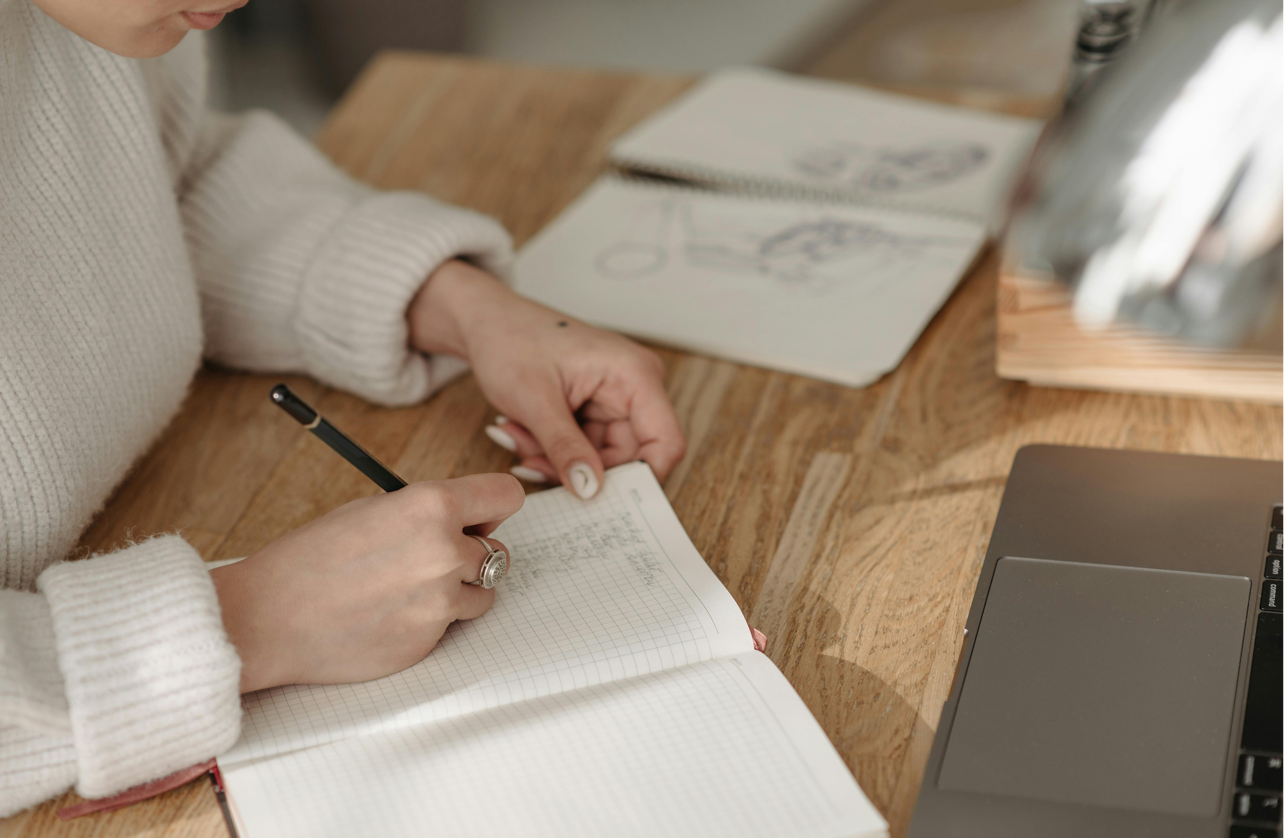 A person in a white sweater writing in a graph paper notebook with a black pen, while another person points at the notebook with a finger. On the table, there is a laptop and some sketches or notes.
