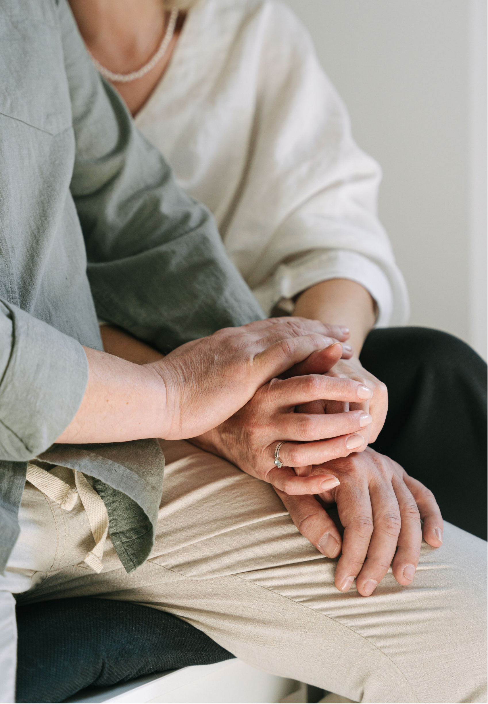 Two people are holding hands, with the focus on their hands and arms. One person has a diamond ring on their finger and appears to be older, while the other is younger. The background is neutral, and they are seated close together.