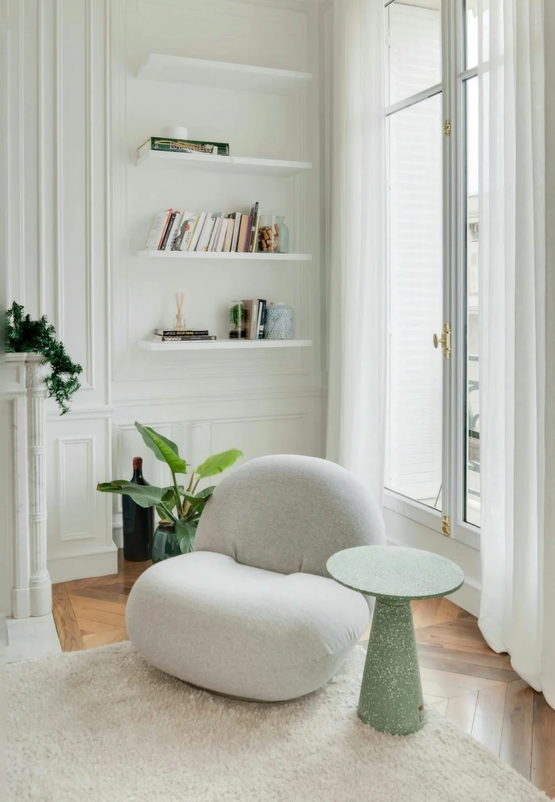 A cozy reading nook with a light gray modern armchair, a small matching side table, a potted plant, white built-in shelves with books, and large window with white curtains letting in natural light in a bright, white-walled room.