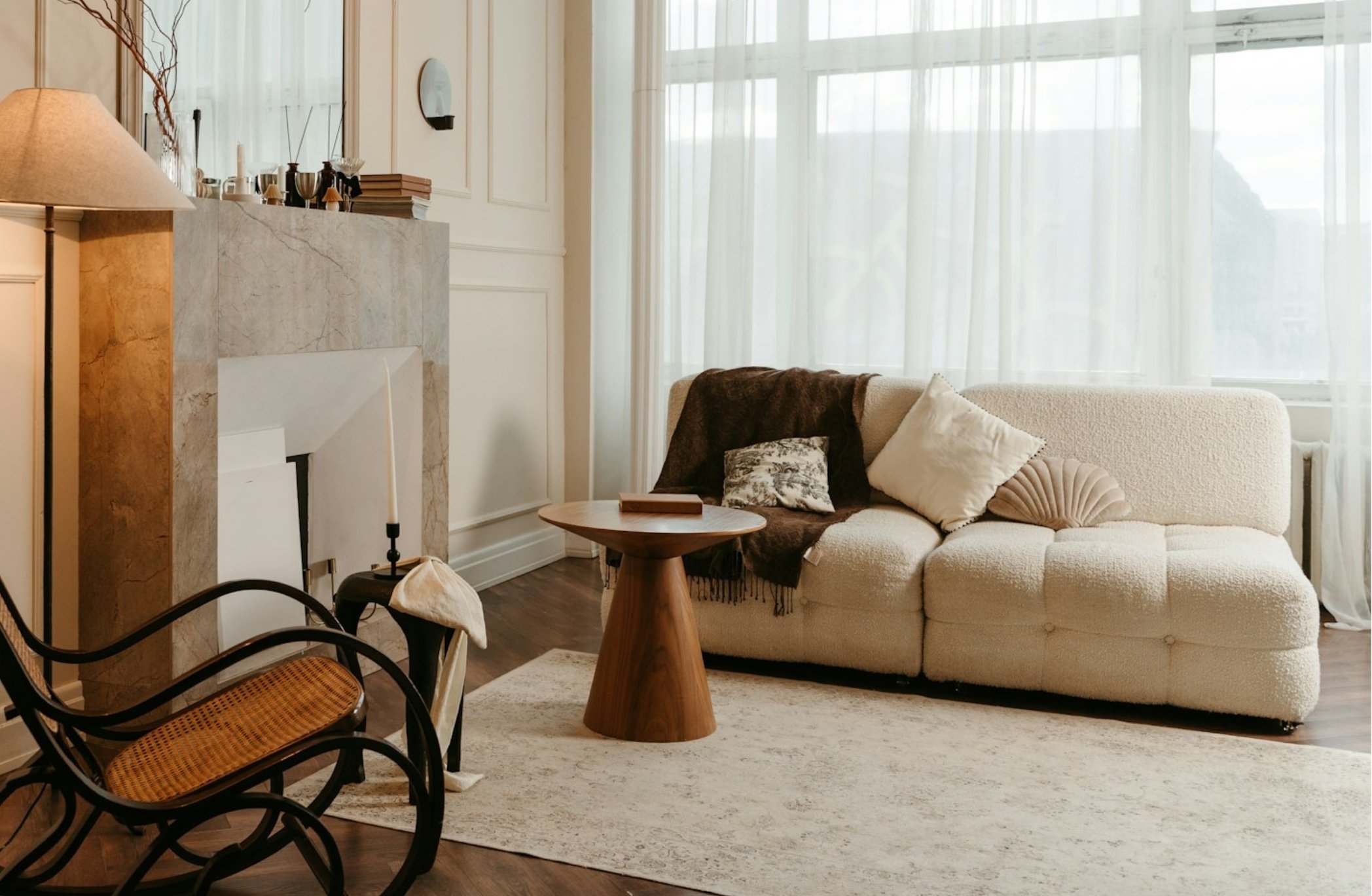 A cozy living room with a cream-colored sofa, decorative pillows, a wooden coffee table, a cream rug, large windows with sheer curtains, a fireplace with a marble mantel, a table with books and decorations, and a black chair with a woven seat.