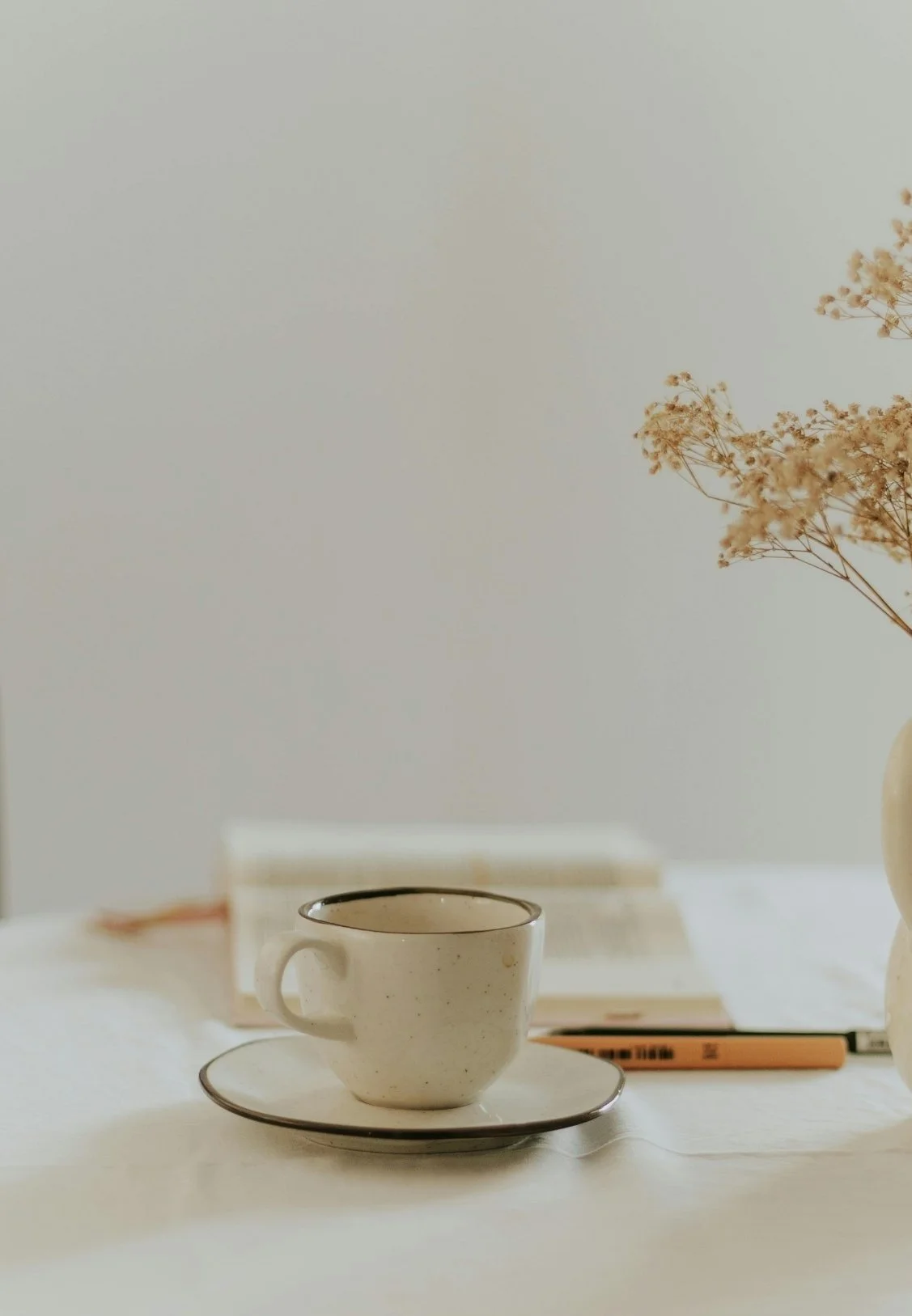 A beige ceramic cup and saucer on a white table, with books and a vase of dried flowers in the background.