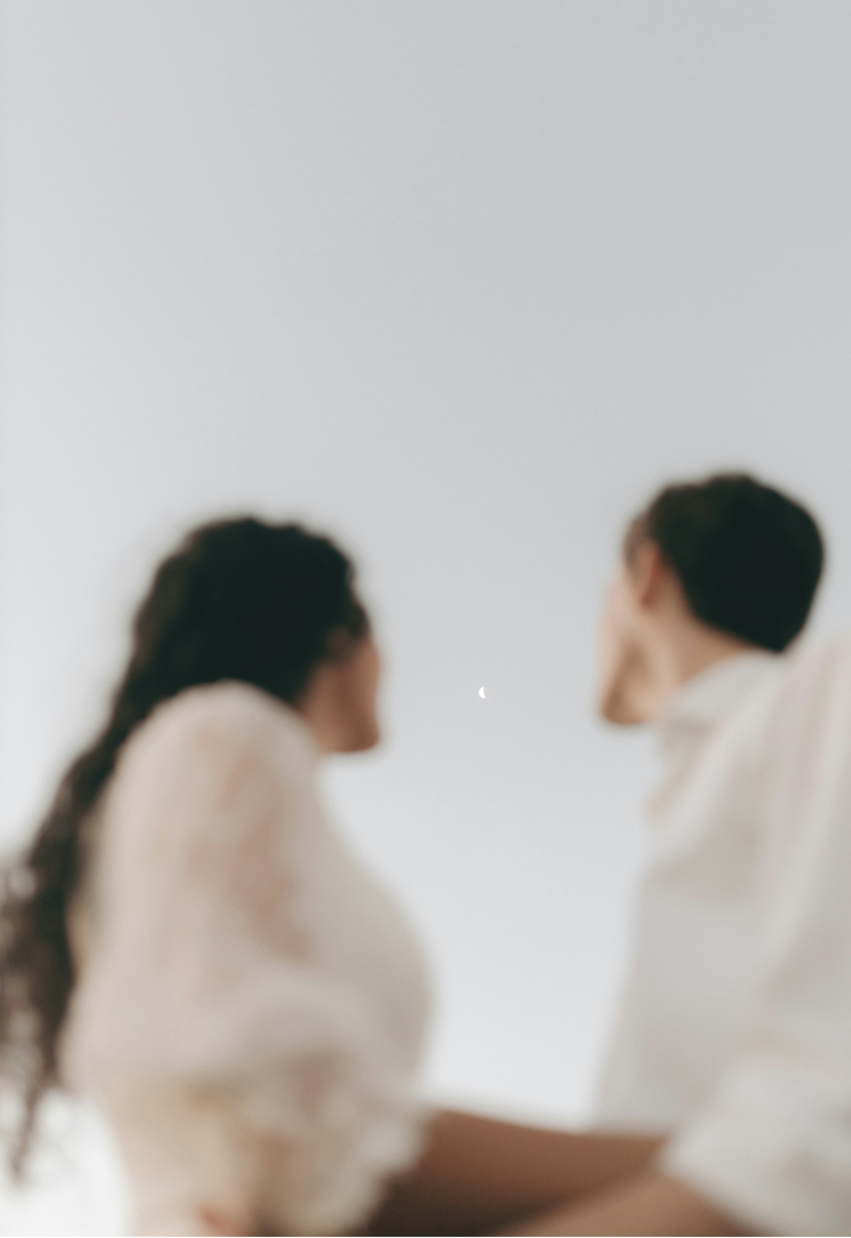 Blurred image of two people, a woman on the left with long hair and a man on the right with short hair, in a minimalistic room with a white wall and a small moon visible in the background.