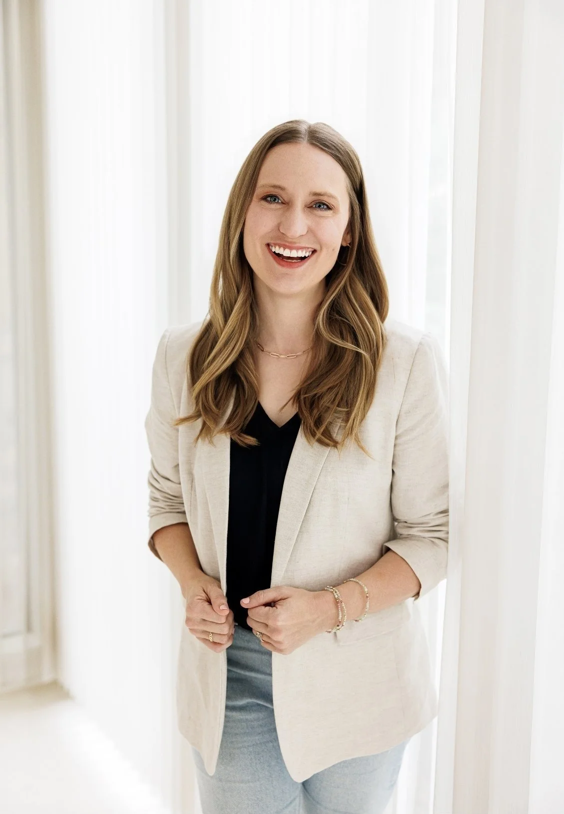 Dr. Laura McKee, Christian counseling in Plano, TX, with long, wavy, light brown hair, wearing a light beige blazer over a black top, standing in front of a bright, white background with sheer curtains.