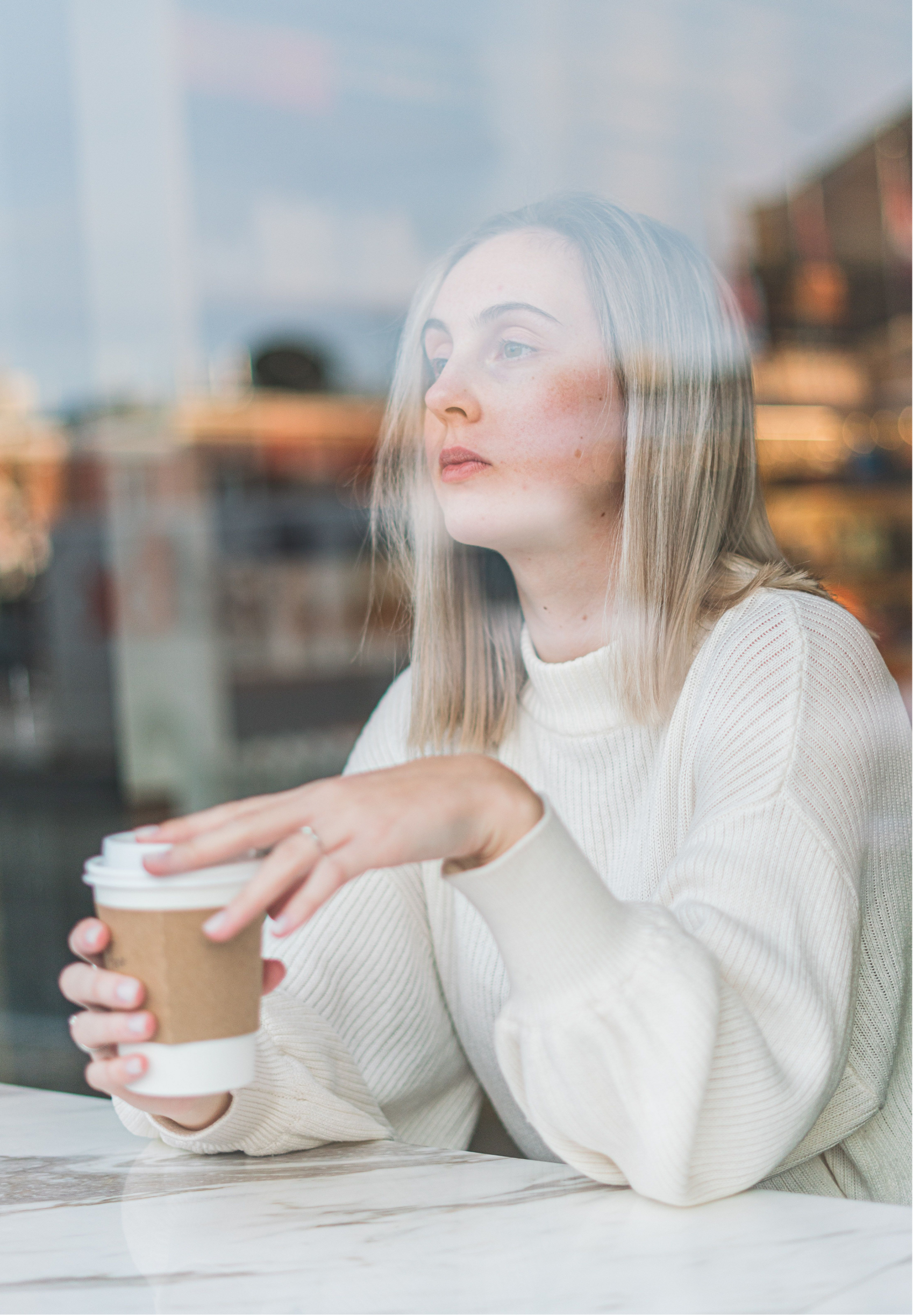 A woman with long blonde hair and freckles, wearing a white sweater, sitting at a table with a marble surface, holding a disposable coffee cup with a cardboard sleeve, in a cafe with blurred background.