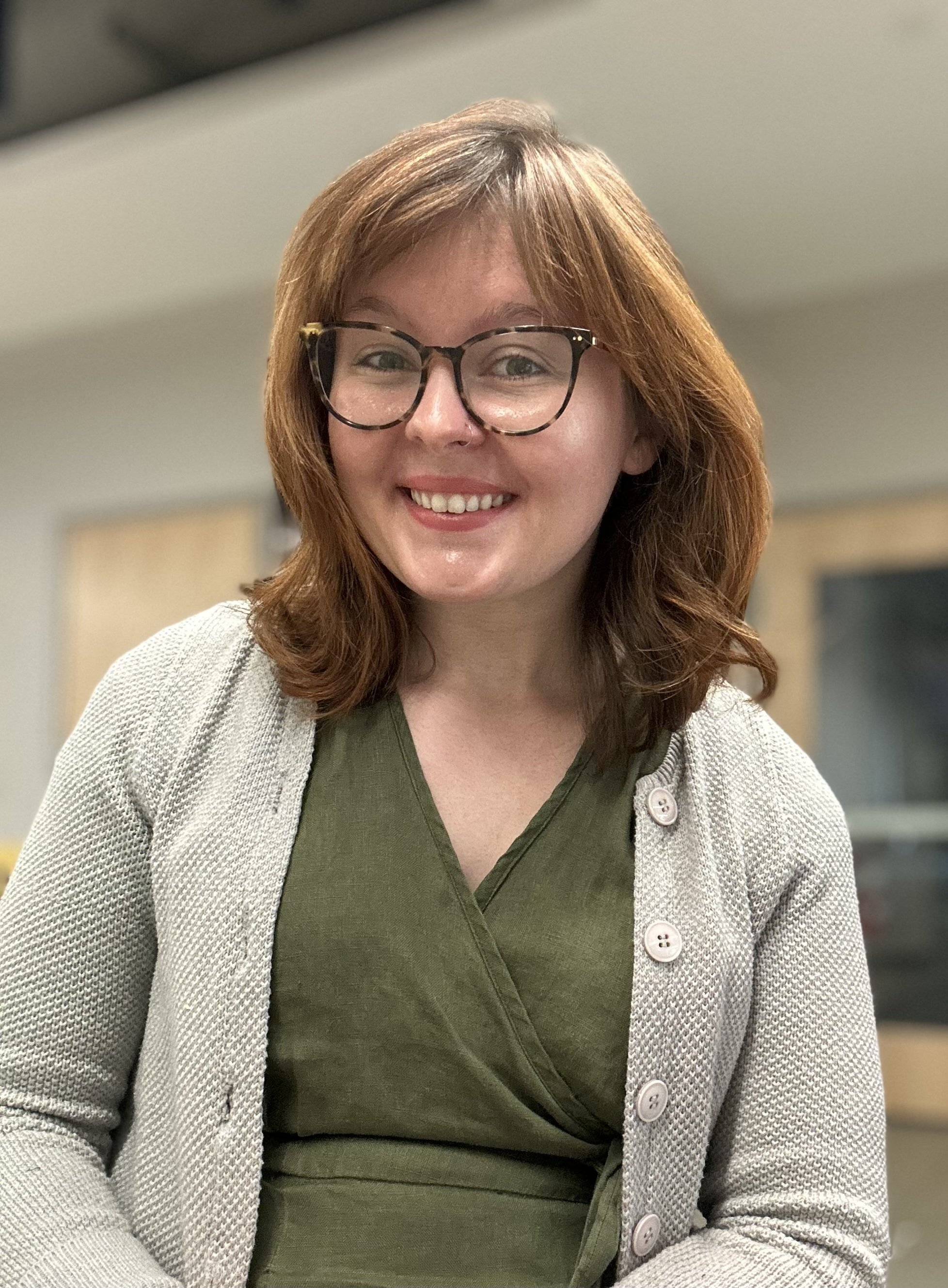 A white cis-gender woman with brown hair smiles, wearing a green shirt and a gray cardigan.