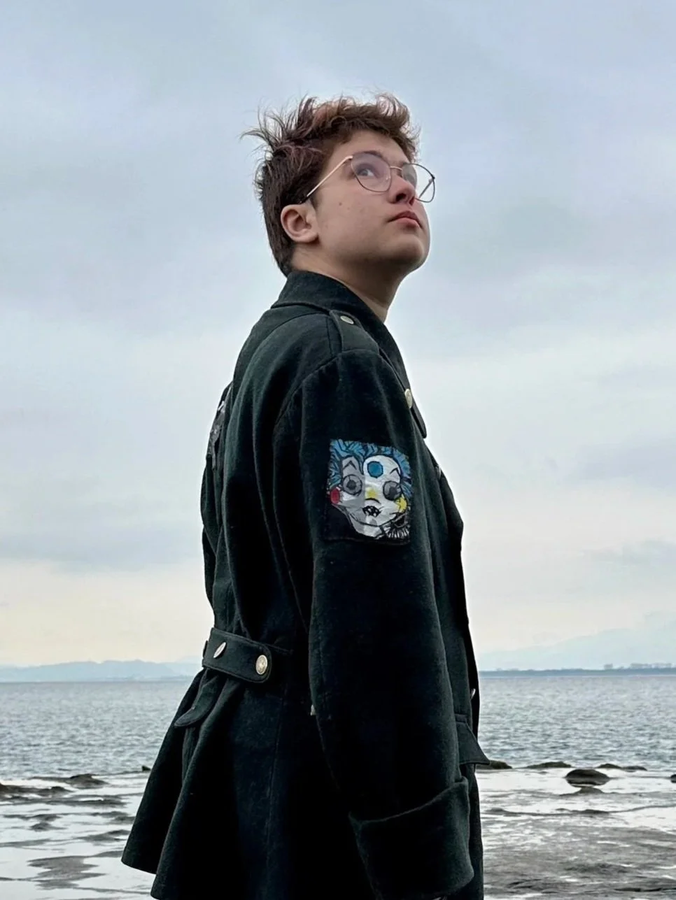 A white non-binary person wears a dark jacket with patches. They look over their shoulder as they stand by a large body of water.
