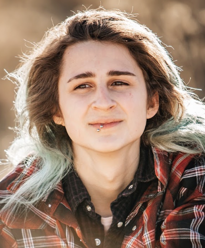 A white trans-fem person with long brown hair that is bleached on the end wears two plaid shirts and a lip ring.