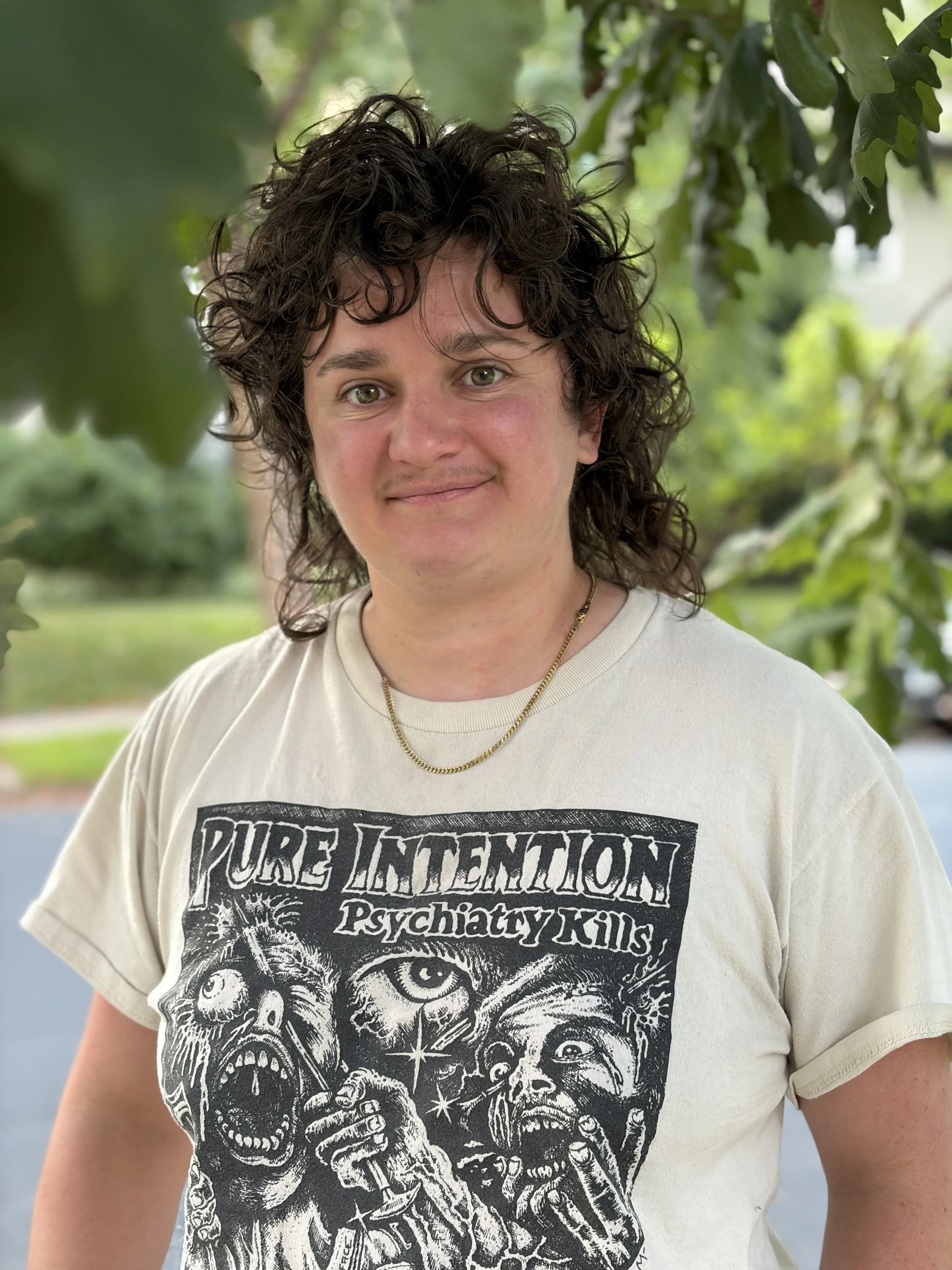 A white non-binary person smiles under a tree. They have dark hair and wear a band t-shirt and a gold chain.