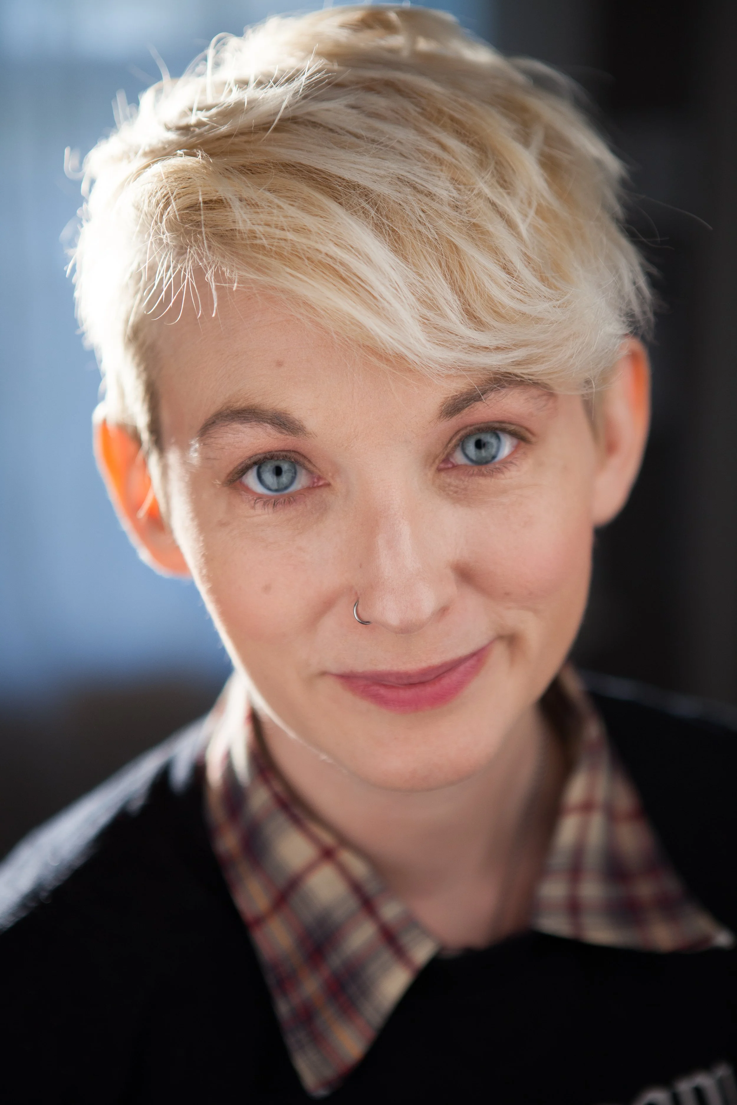 A white non-binary person with blonde hair and a nose ring smiles, wearing a plaid collared shirt under a dark sweater.