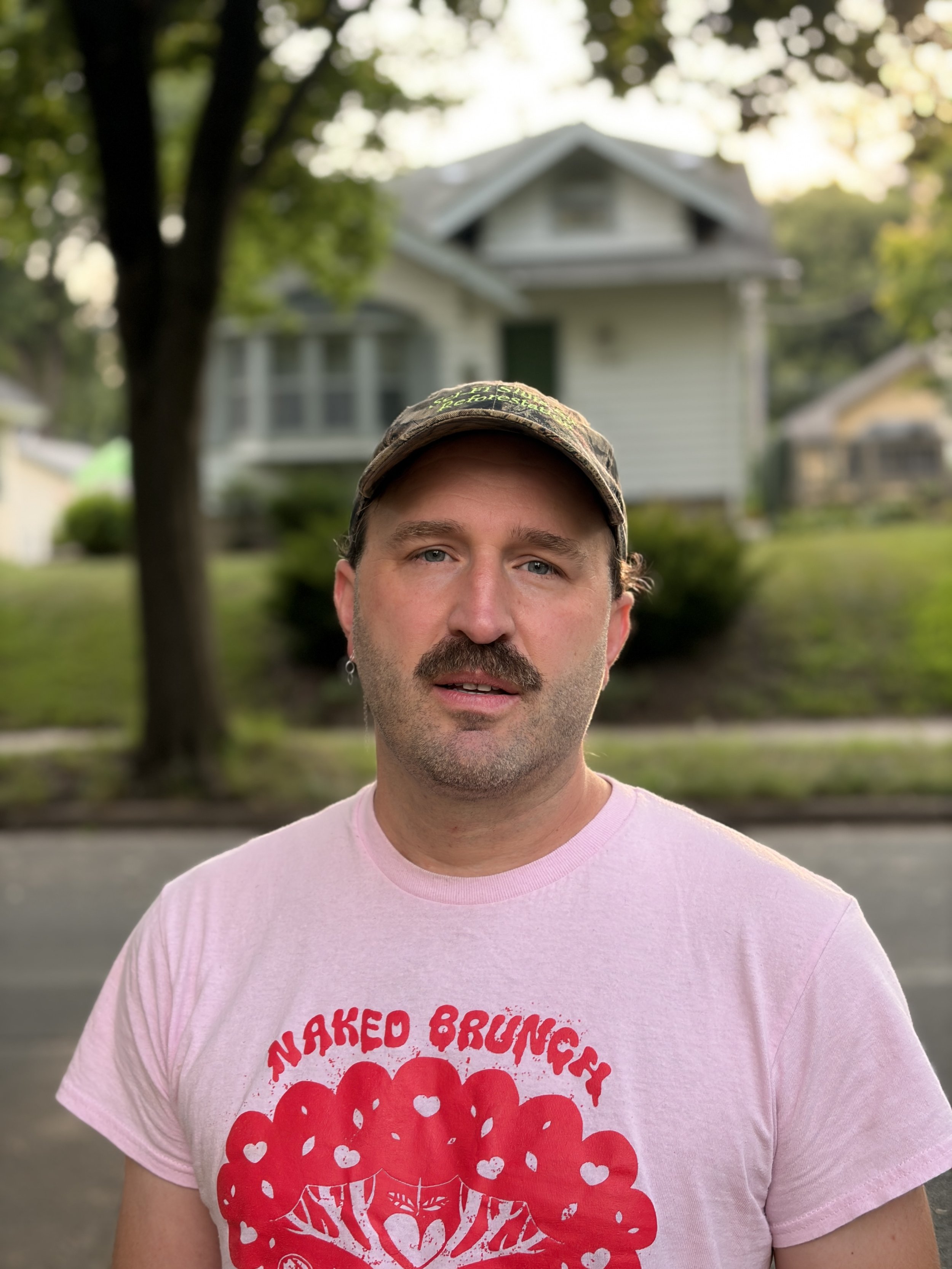 A white cis-gender man with a mustache standing in front of a wooded neighborhood wears a pink band t-shirt and a camp hat.