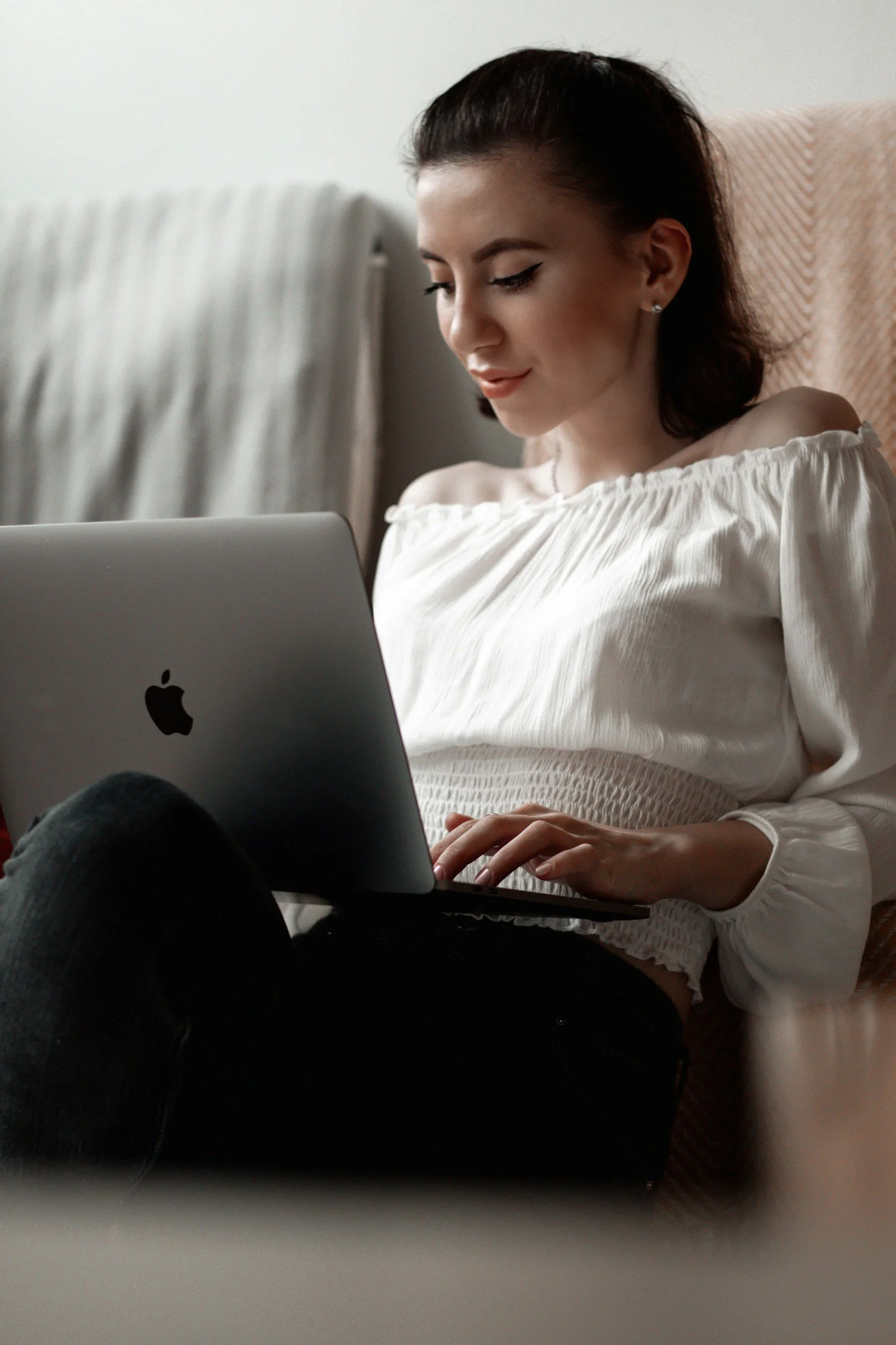 A young woman with dark hair and winged eyeliner is sitting on a beige cushioned chair, looking at a silver MacBook laptop on her lap, smiling gently. She is wearing an off-shoulder white blouse with ruffled edges.