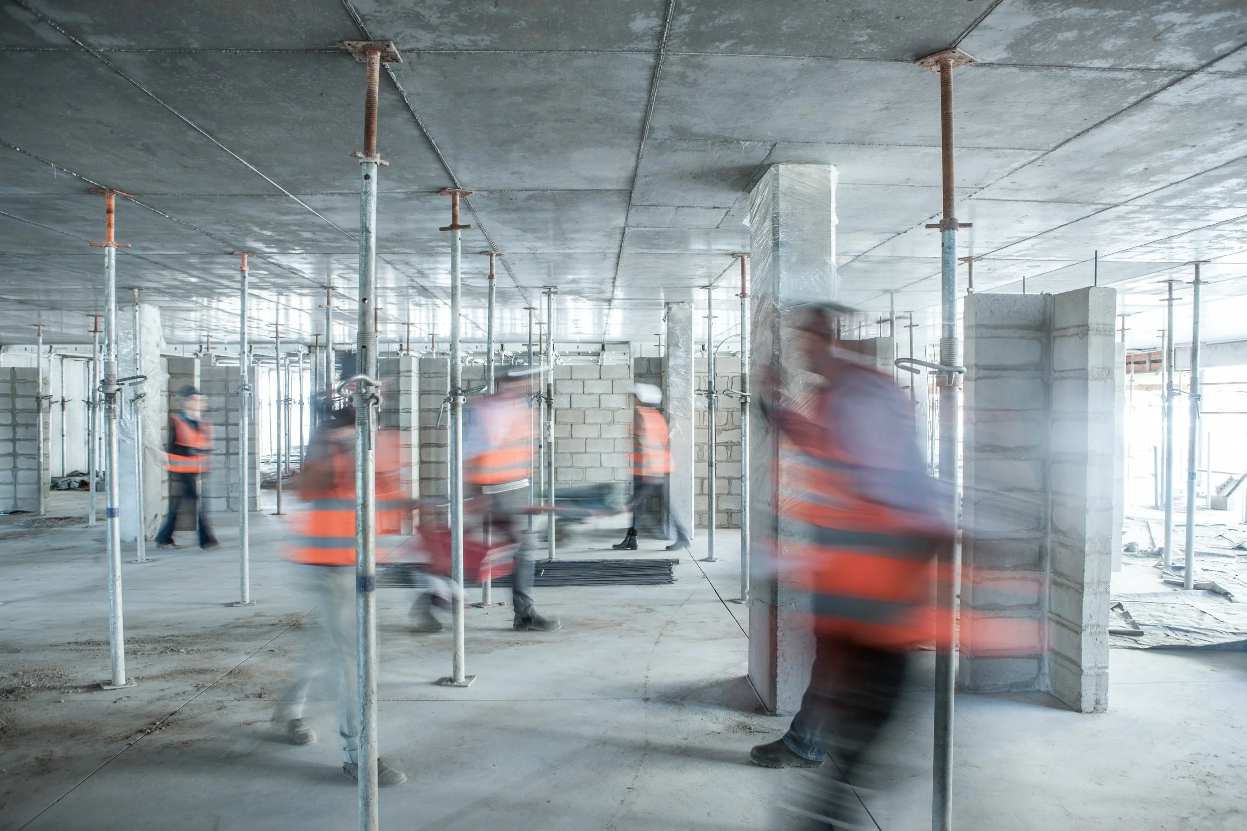 Construction workers in orange vests working inside a building under construction with metal scaffolding and unfinished concrete walls.