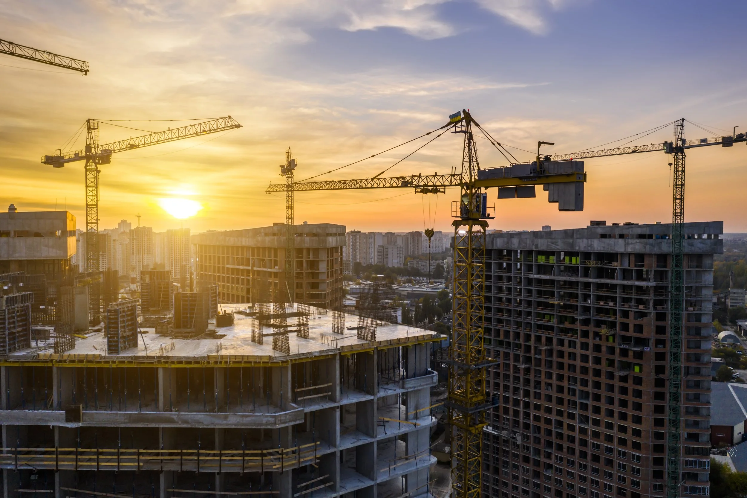 A cityscape at sunset with multiple construction cranes and partially built high-rise buildings.