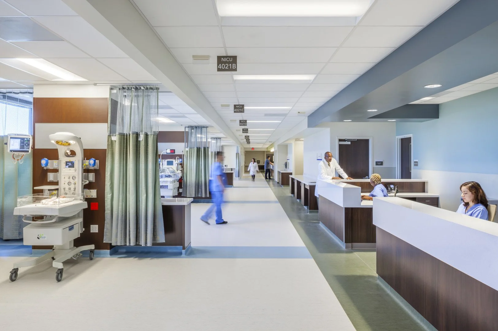 Hospital nursery with medical staff and equipment, NICU signs, infant incubator, nurses working at reception desks, and patients in the background.