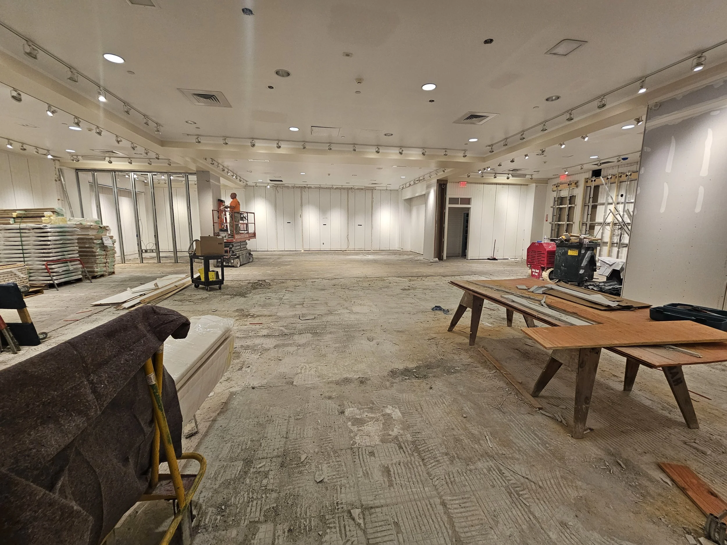 Construction workers installing ceiling panels in a large room, with various construction materials and tools scattered on the unfinished floor.
