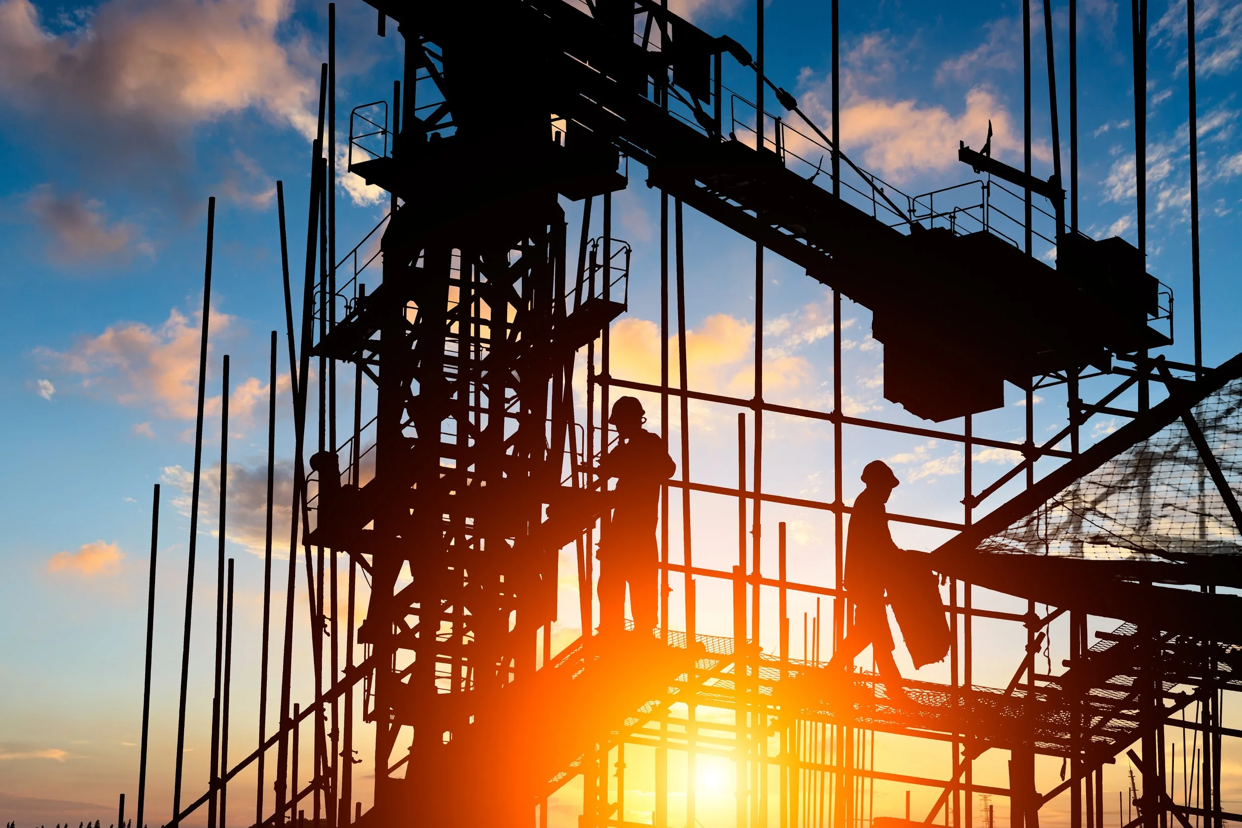 Silhouette of construction workers on scaffolding against a sunset sky with clouds.