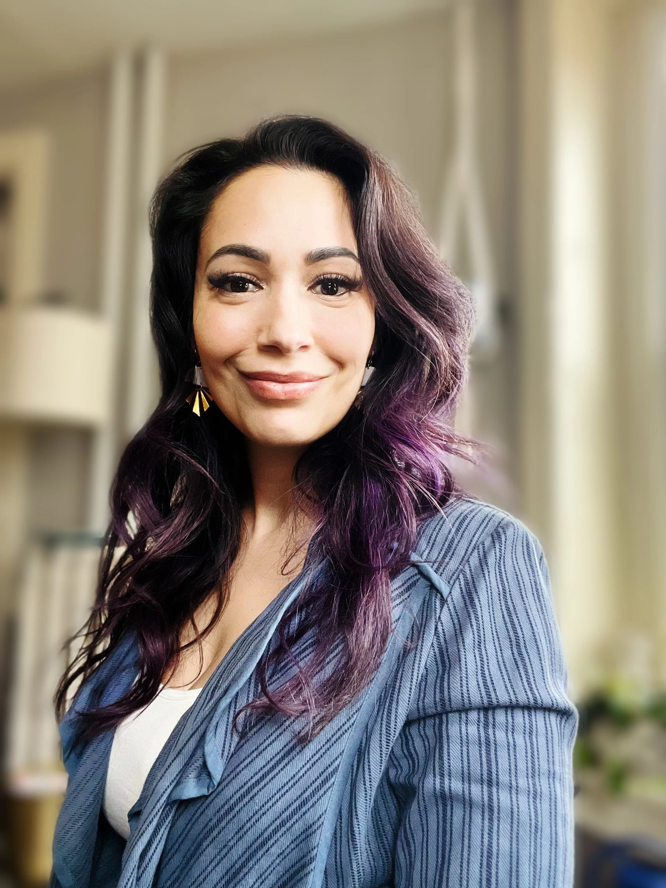A woman with black and purple wavy hair smiling in a room with blurred background.