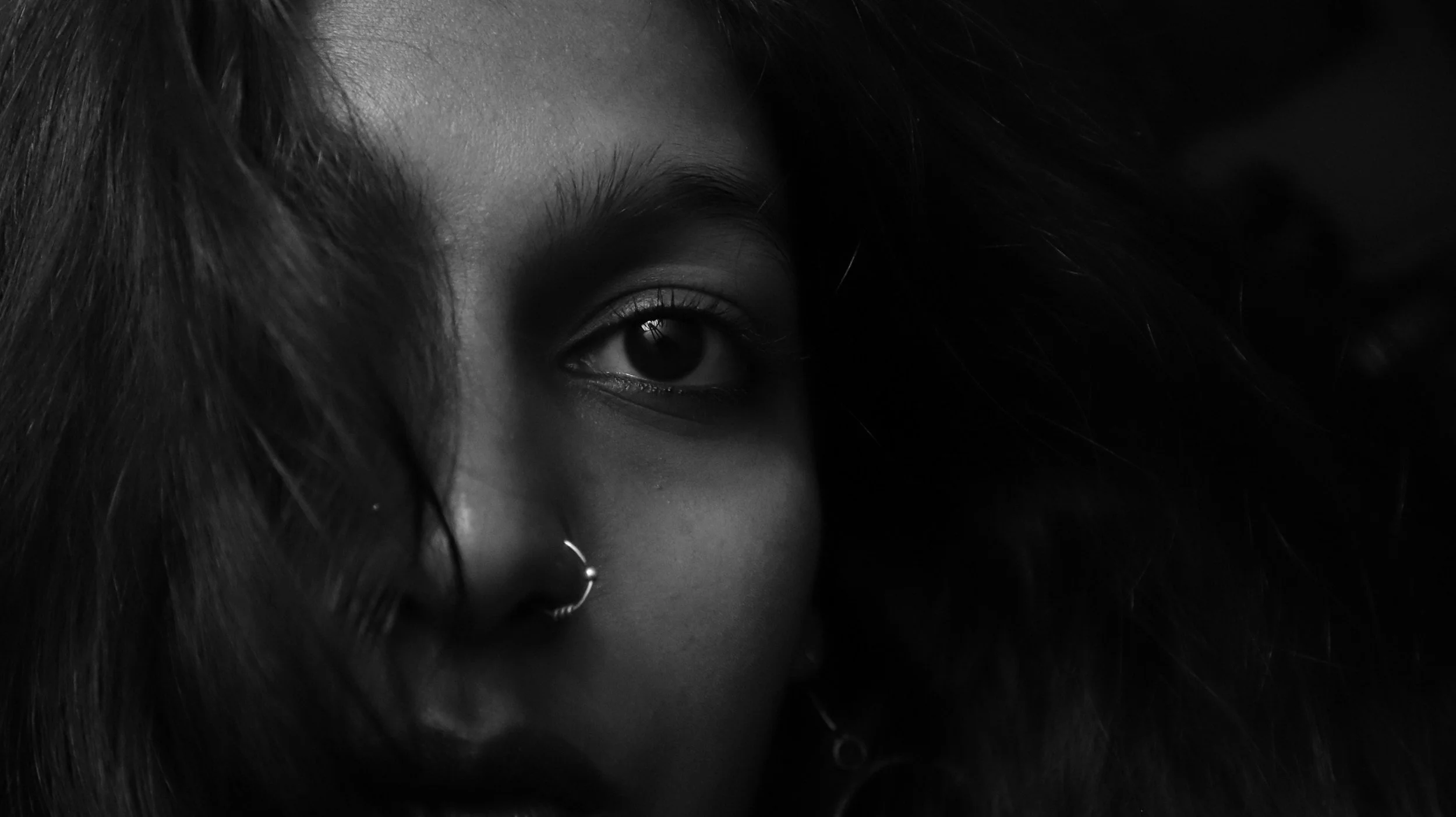 Close-up of a woman's eye with dark hair and a nose piercing, in black and white.