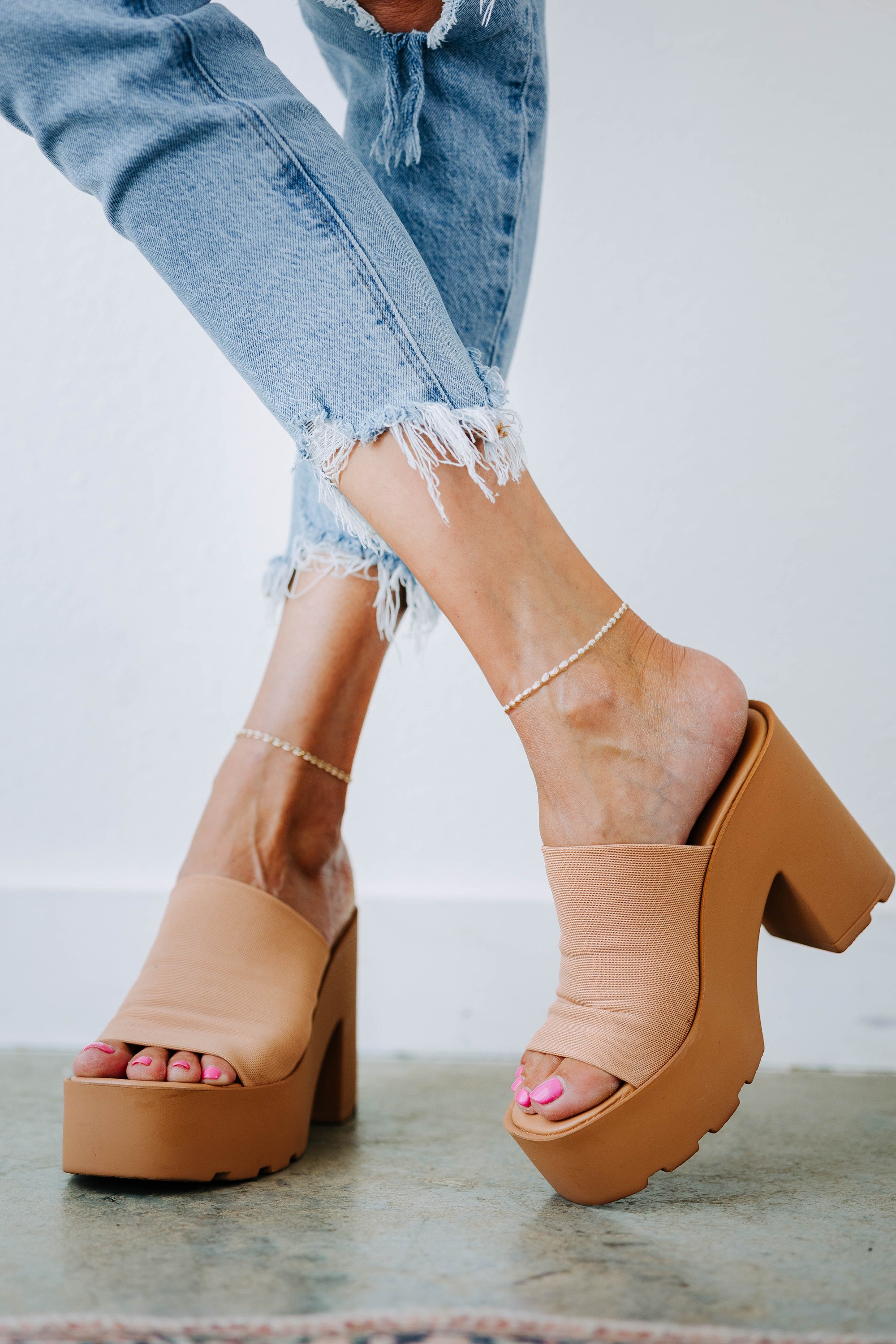 Close-up of a woman's legs wearing distressed blue jeans with frayed hems and beige platform high-heeled sandals with pink toenail polish and ankle jewelry, against a plain white background.