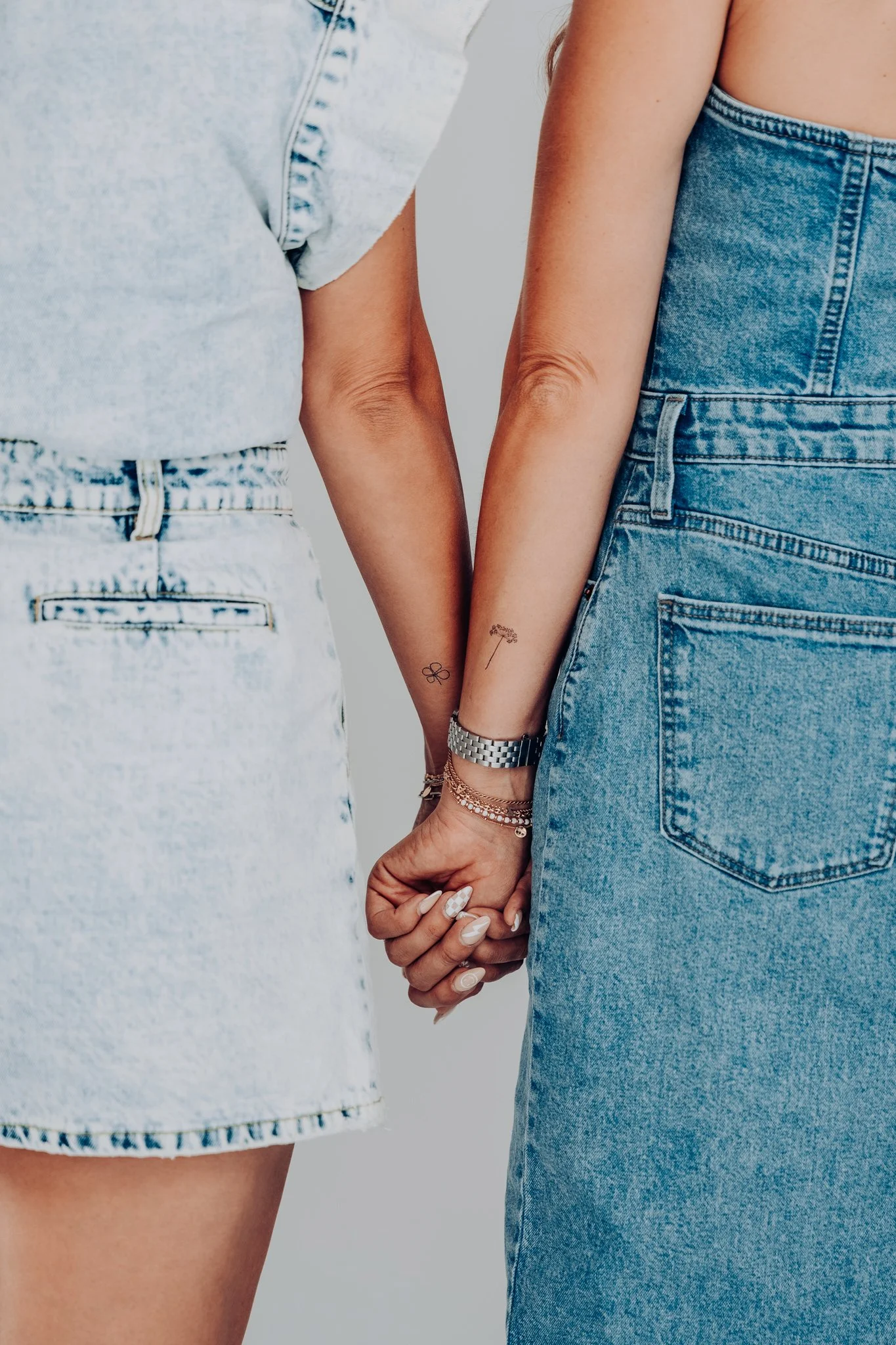 Two women holding hands, one wearing a light-colored denim skirt, the other wearing a denim dress, both with tattoos on their arms and wearing jewelry.